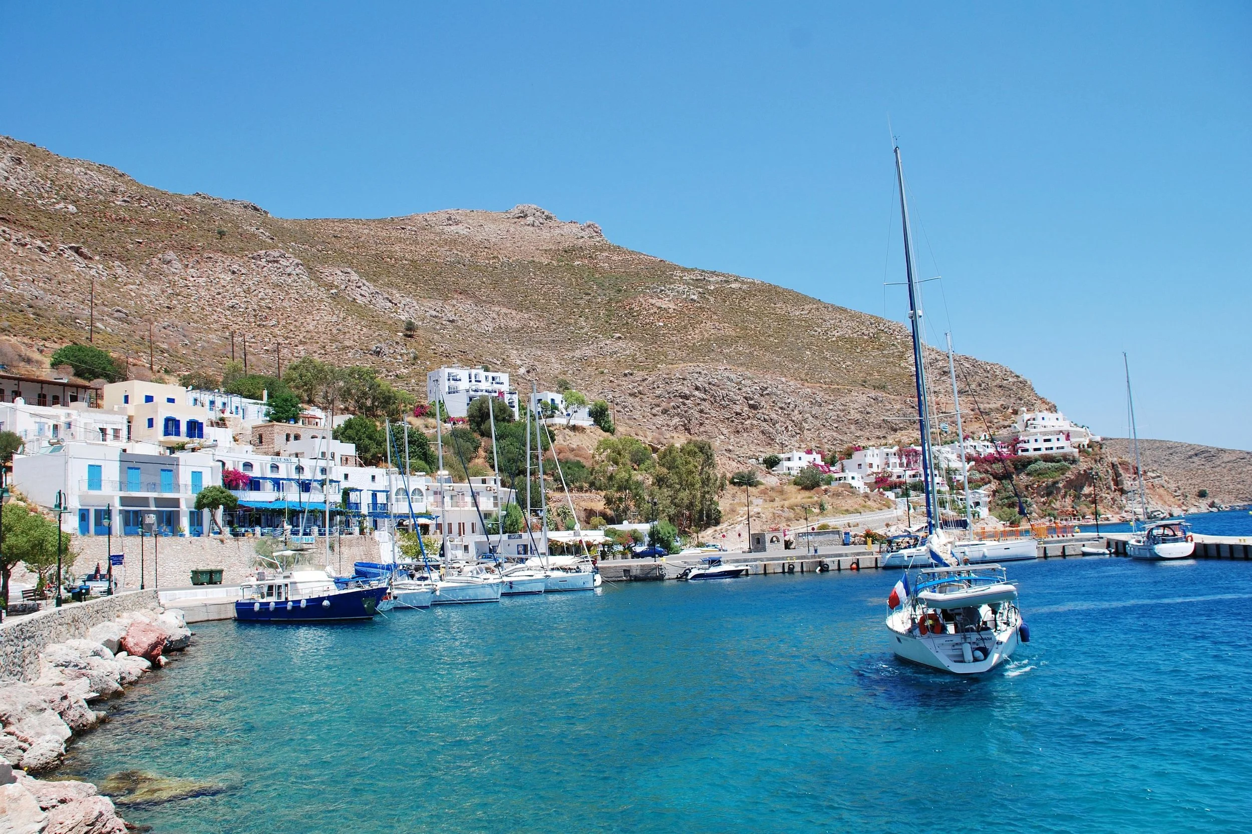 A scenic harbor with several sailboats docked and white buildings with blue accents along the shoreline, set against a hilly, arid landscape under a clear blue sky.