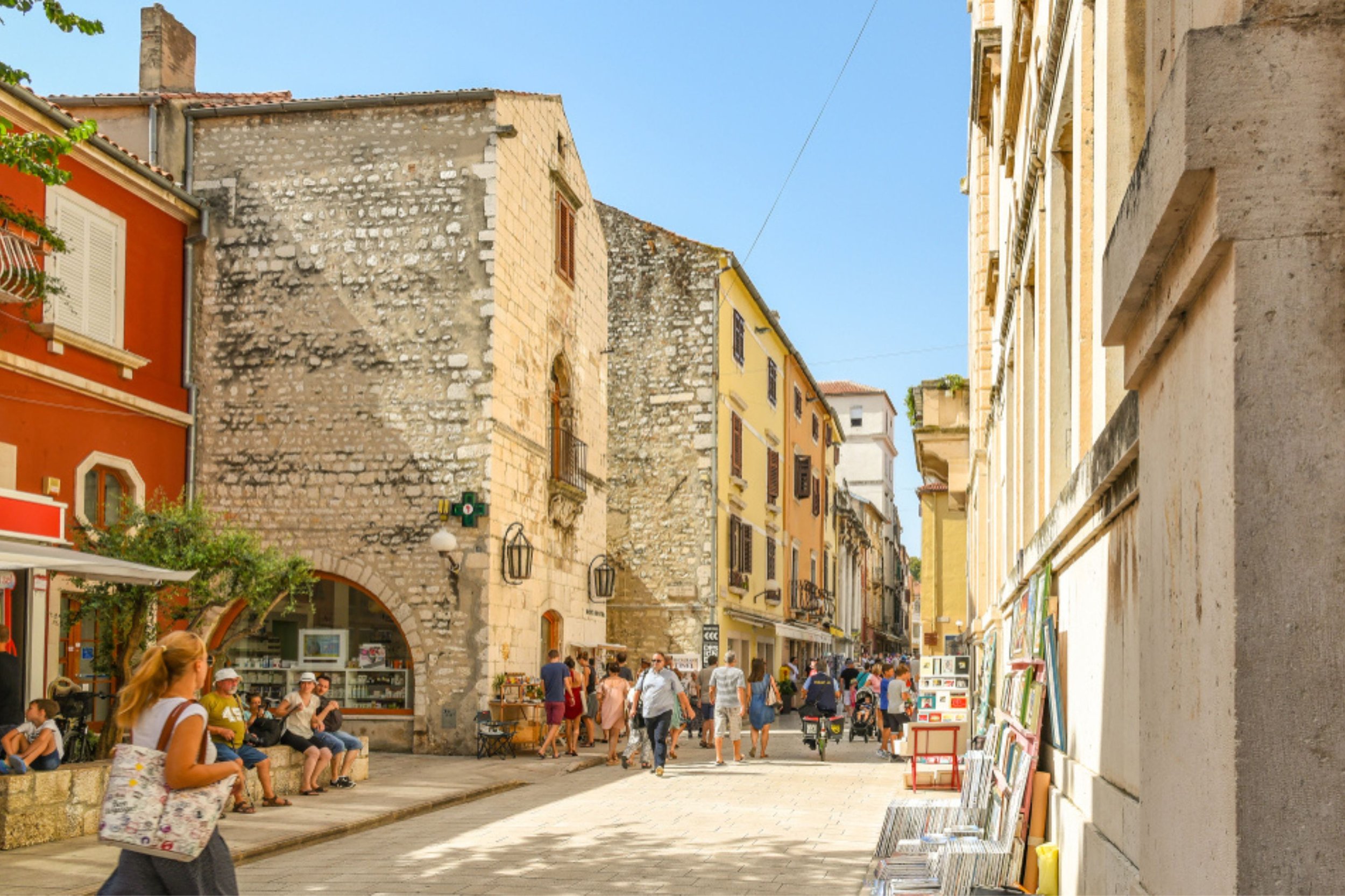 A lively street scene with tourists and locals walking, shopping, and sitting outside cafes in a European town, surrounded by colorful historic buildings under a clear blue sky.