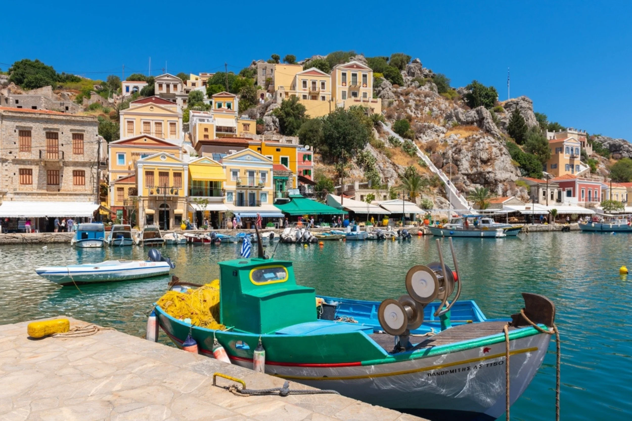 Colorful boats docked in a harbor with pastel buildings and hillside in the background under a clear blue sky.