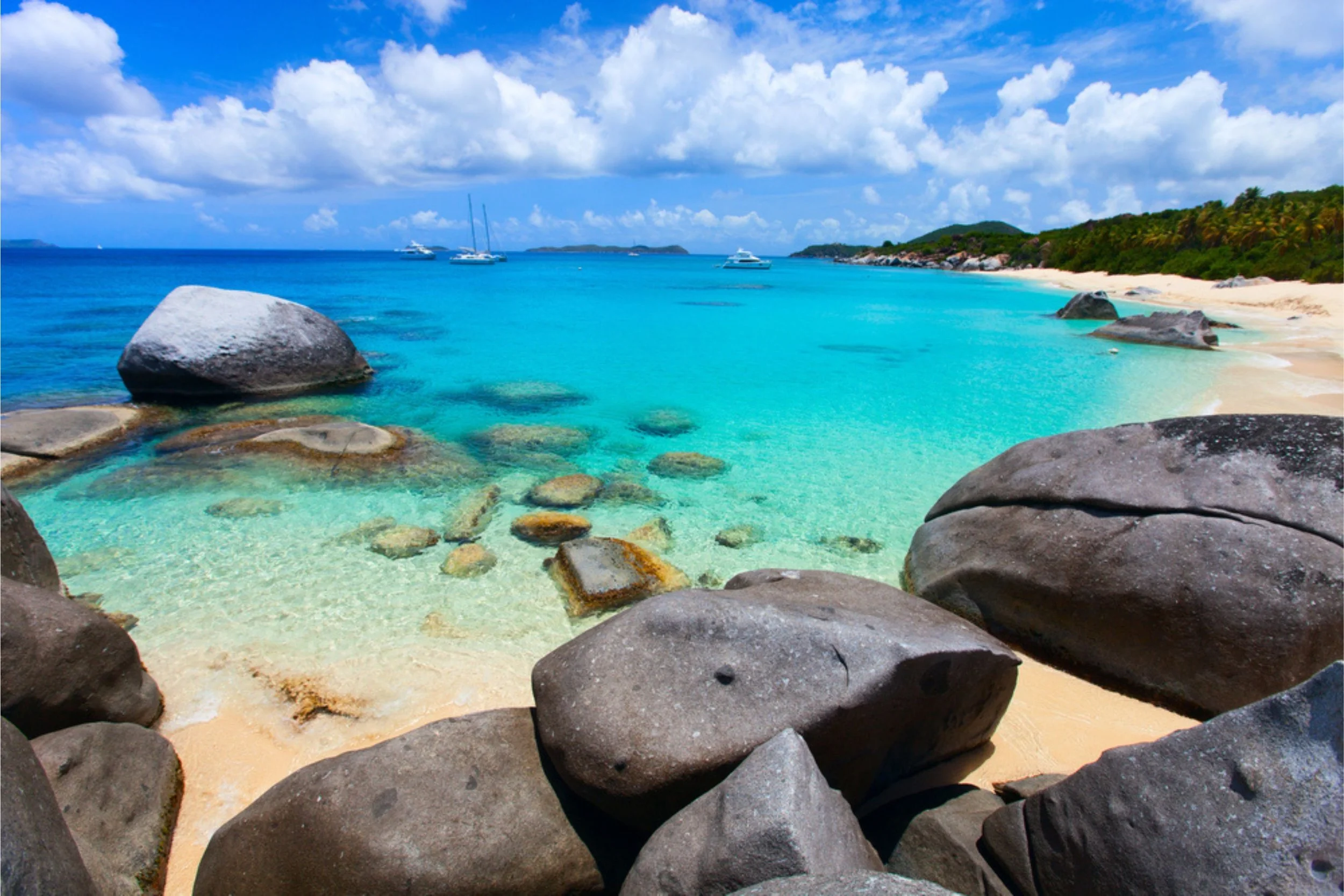 Turquoise ocean water with large rocks along a sandy beach, boats in the distance, green hills, and partly cloudy sky.
