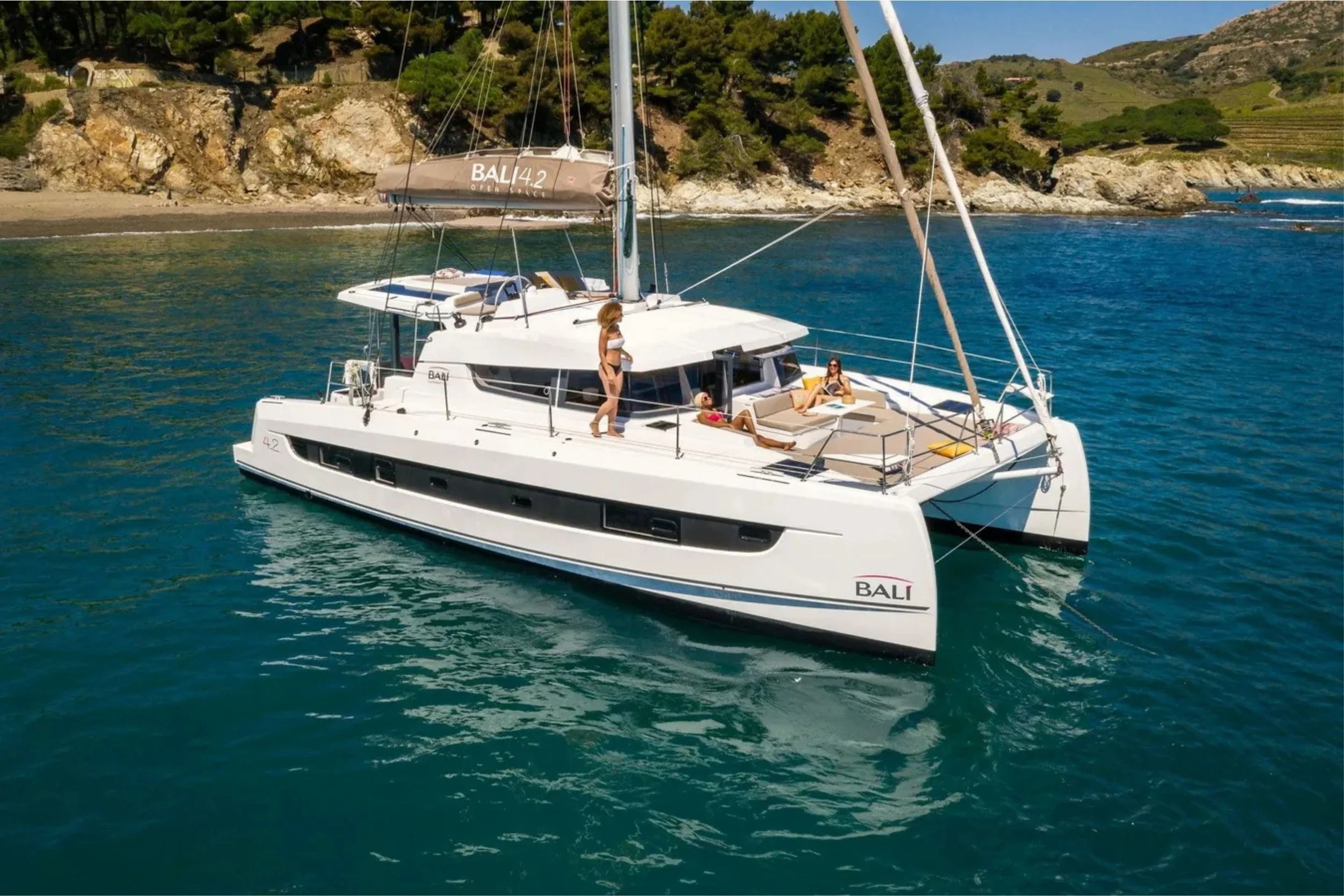 A white sailboat named BALI 4.2 anchored near a rocky shoreline with green trees, with two women relaxing on the deck and one woman standing, enjoying sunny weather.