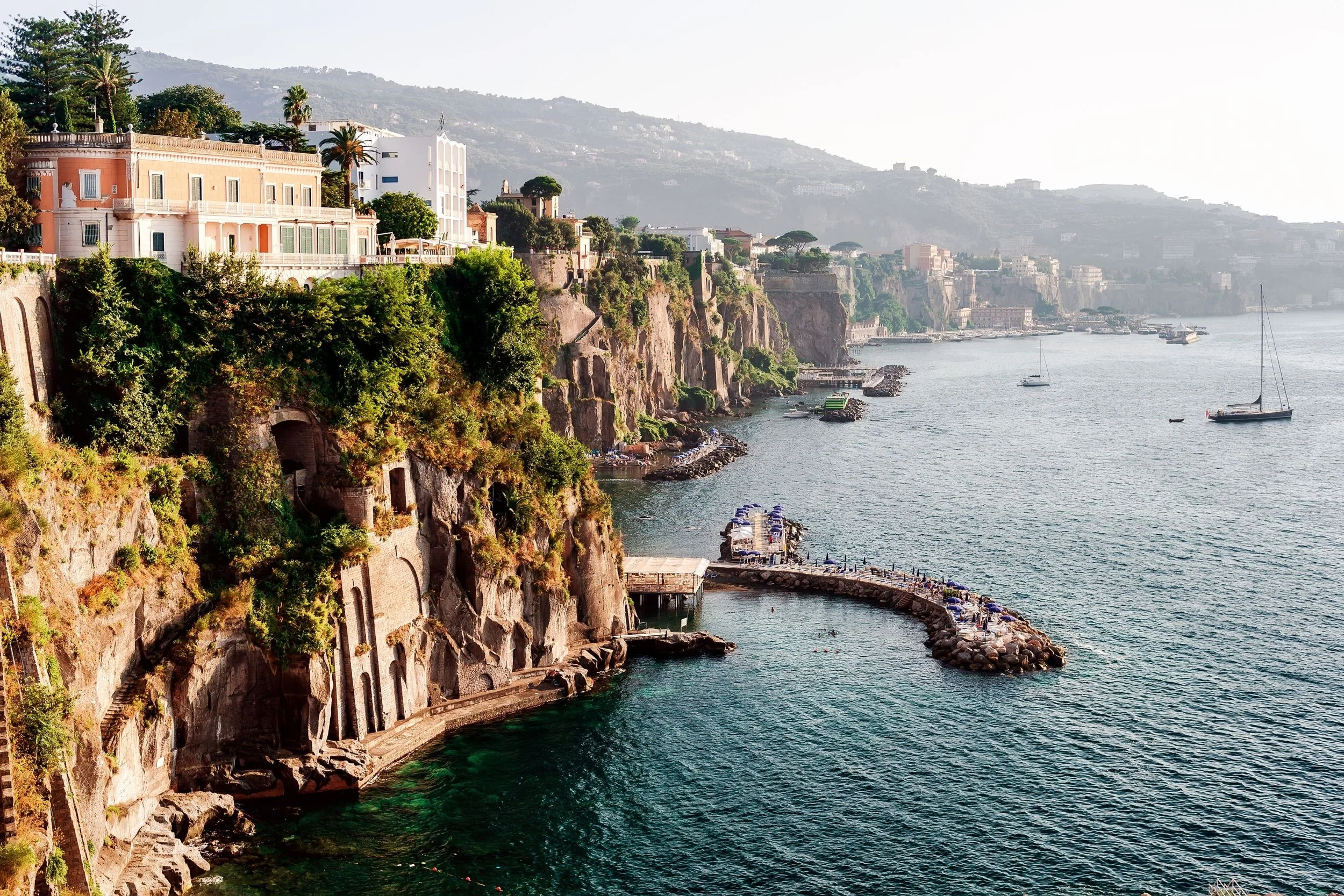 Coastal scene with cliffs, colorful buildings, boats, and a harbor along the water with hills in the background.