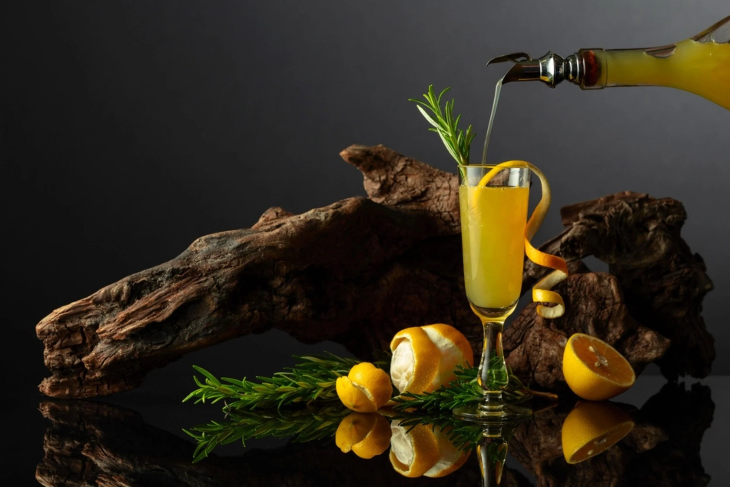 A cocktail in a tall glass with lemon peel and rosemary, topped with a lemon twist, being poured from a bottle, with lemons, lemon peel, and rosemary arranged on a reflective surface against a dark background.