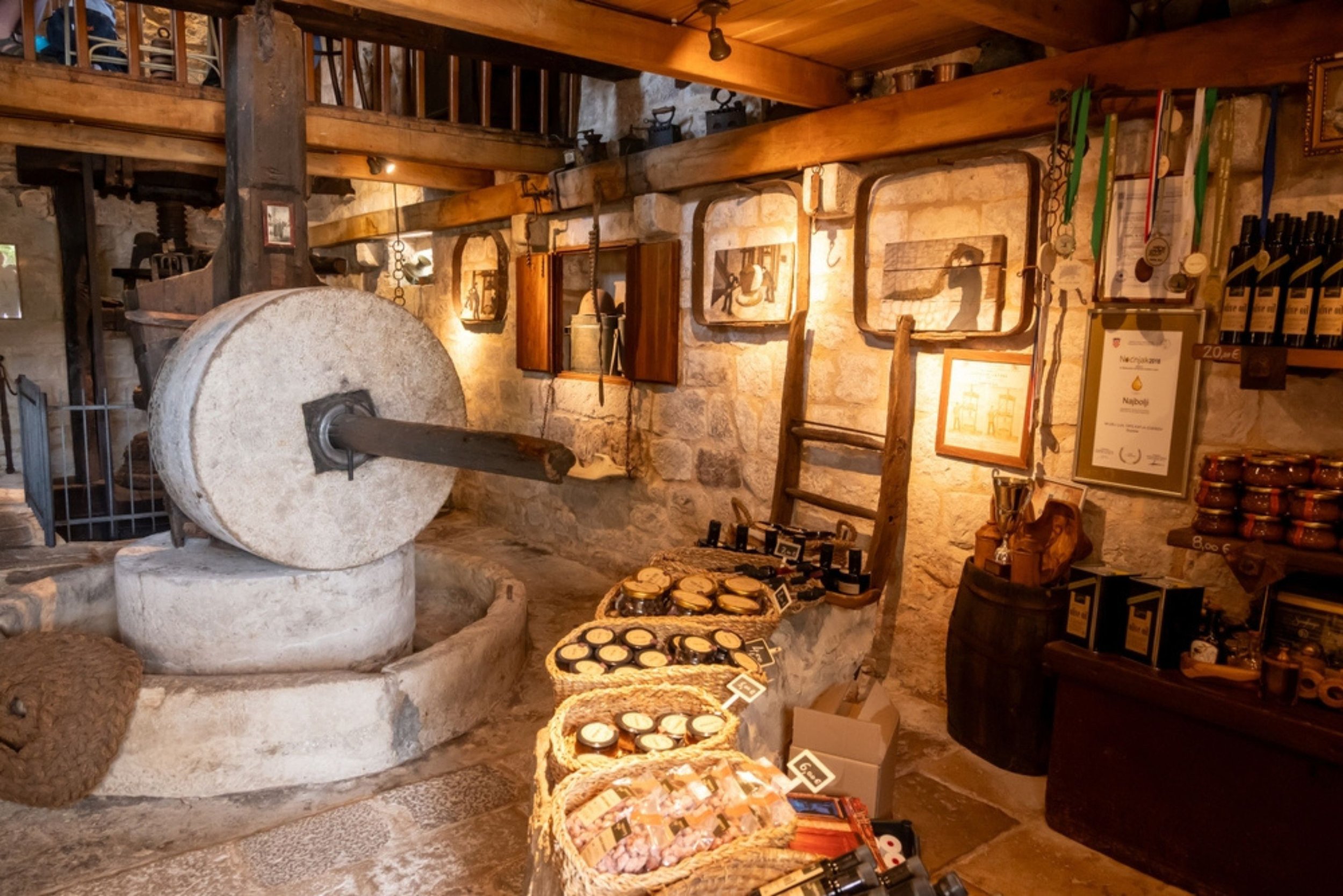 A rustic store interior featuring a stone mill, wooden beams, framed artwork on stone walls, shelves with jars and bottles, and baskets of various products for sale.