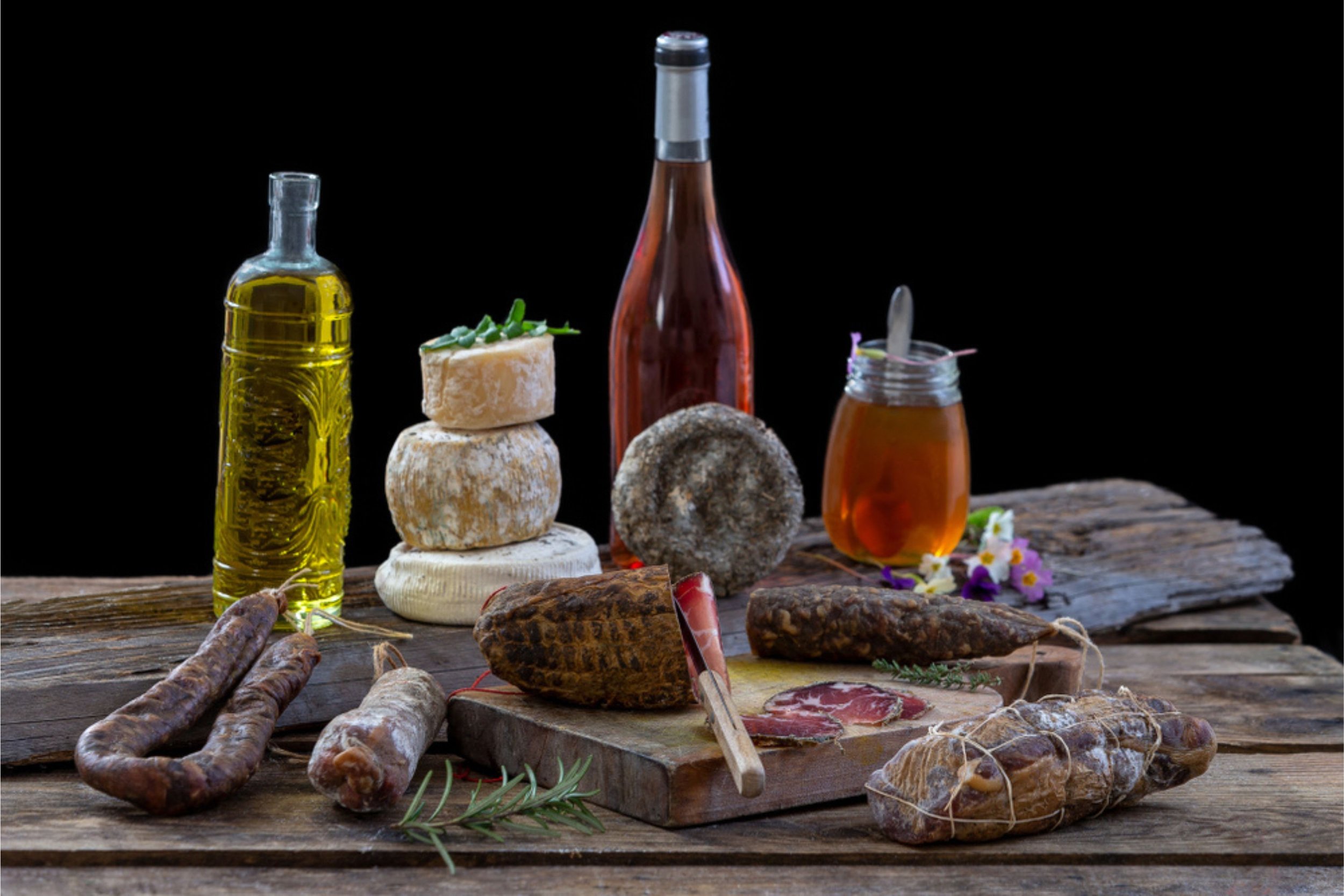 Assorted cheeses, smoked meats, sausages, herbs, wine, and honey on a rustic wooden table against a black background.