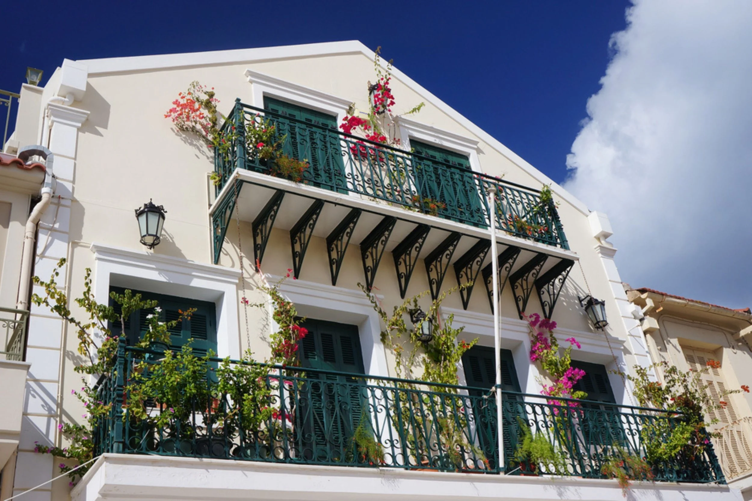 A white multi-story building with green doors and balconies decorated with flowers and plants, against a blue sky with some clouds.