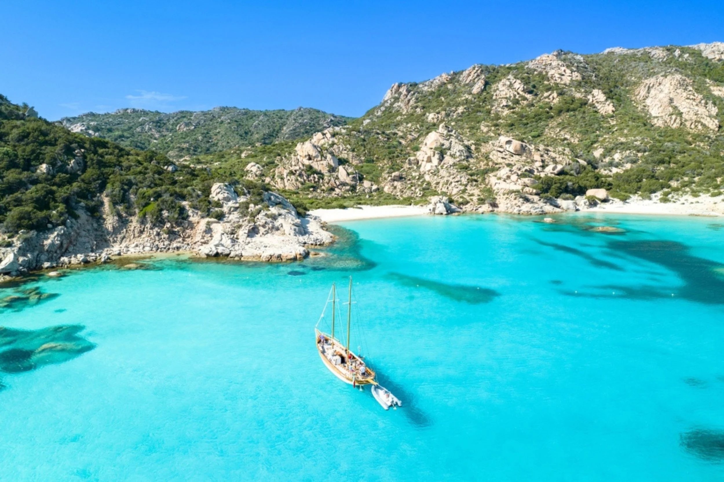 A boat floating on clear turquoise water near rocky shoreline and lush green hills under a bright blue sky.