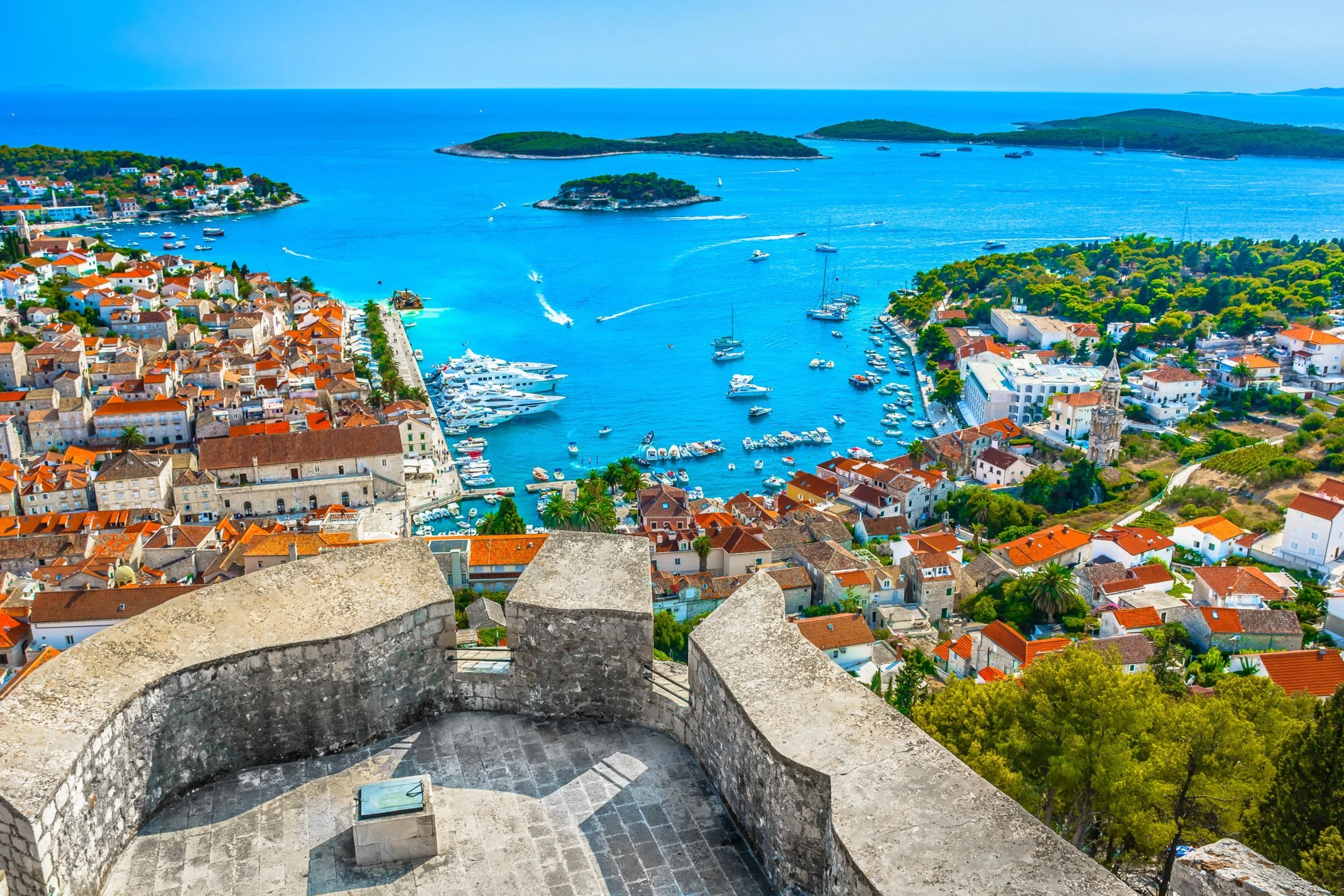A scenic coastal town viewed from a high vantage point showing red-tiled roofs, a marina with boats, and the blue sea with small islands in the background.