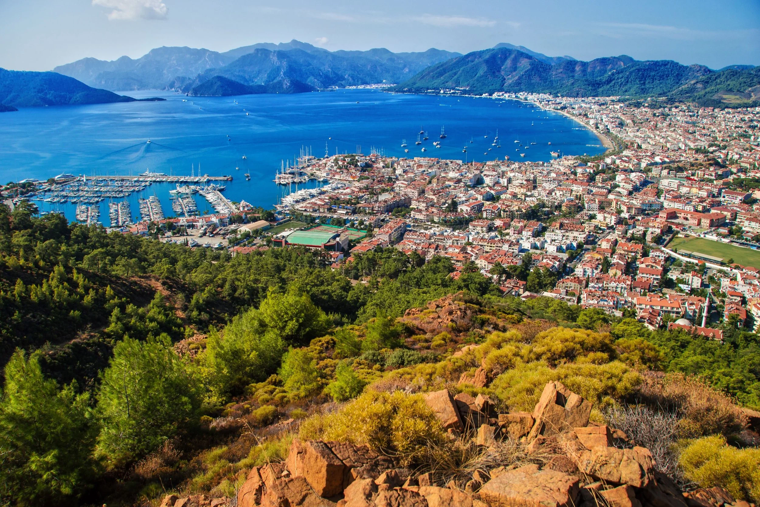 Aerial view of a coastal city with a large harbor filled with numerous boats and yachts, surrounded by green mountains and a densely built urban area.
