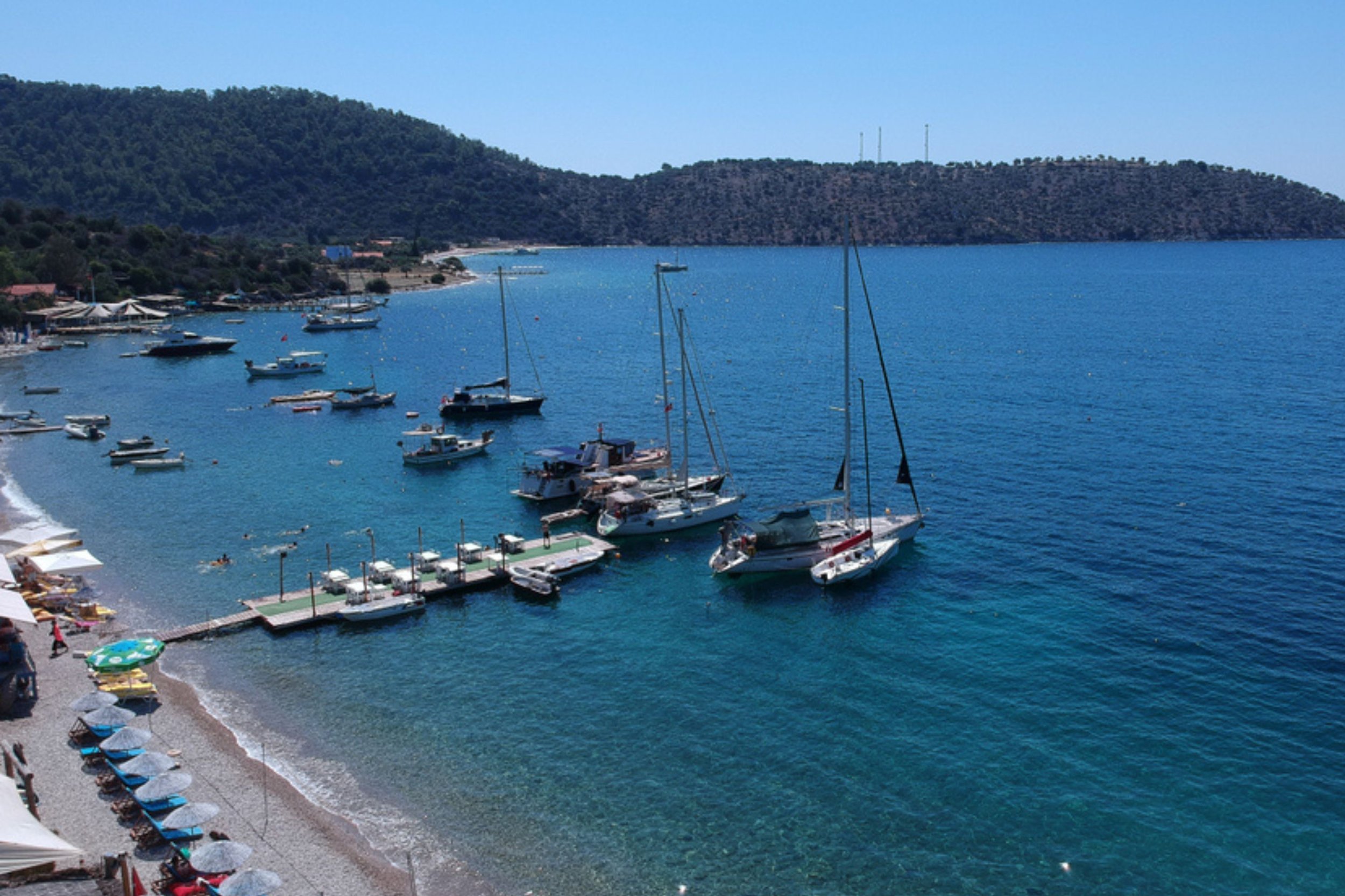 A marina with sailboats and motorboats docked by a pebble beach. The beach has umbrellas and lounge chairs, and the coastline is lined with trees and hills under a blue sky.