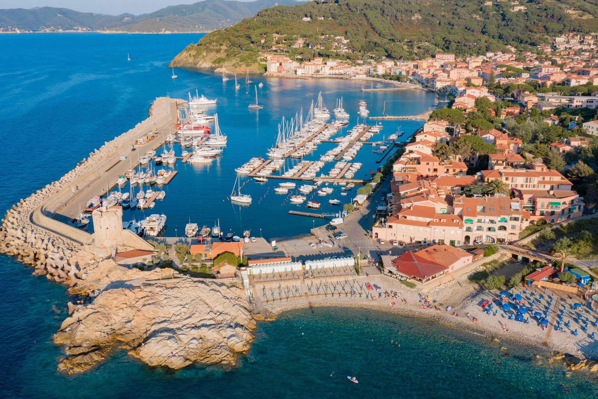 Aerial view of a marina with boats, a small beach with umbrellas, and a coastal town with orange-roofed buildings on a hillside.
