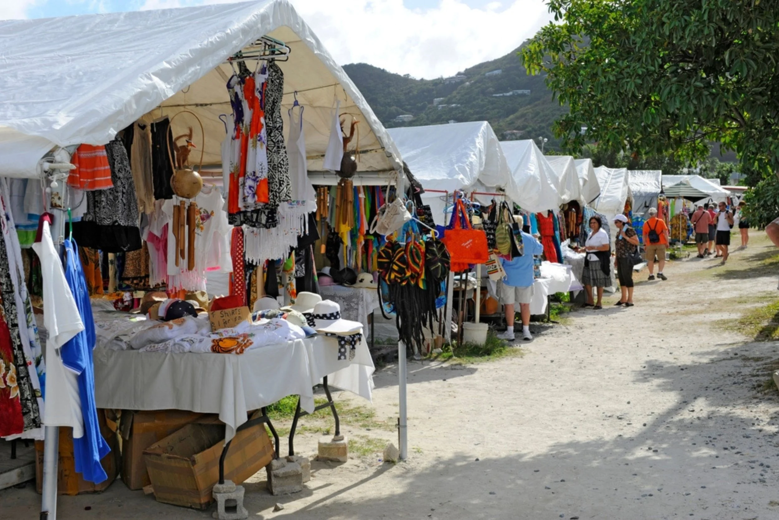 Outdoor market with white tents selling clothes and accessories, people shopping on a dirt path, greenery and mountain in the background.