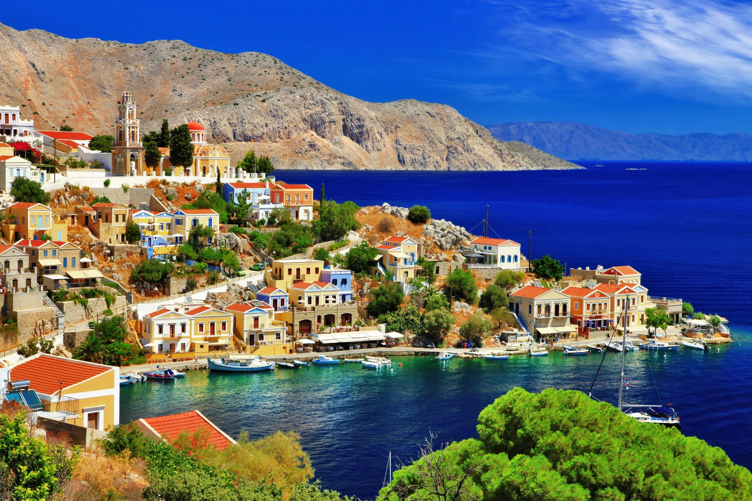 Colorful houses along a hillside next to a deep blue sea with boats docked at the shore, mountainous landscape in the background under a partly cloudy sky.