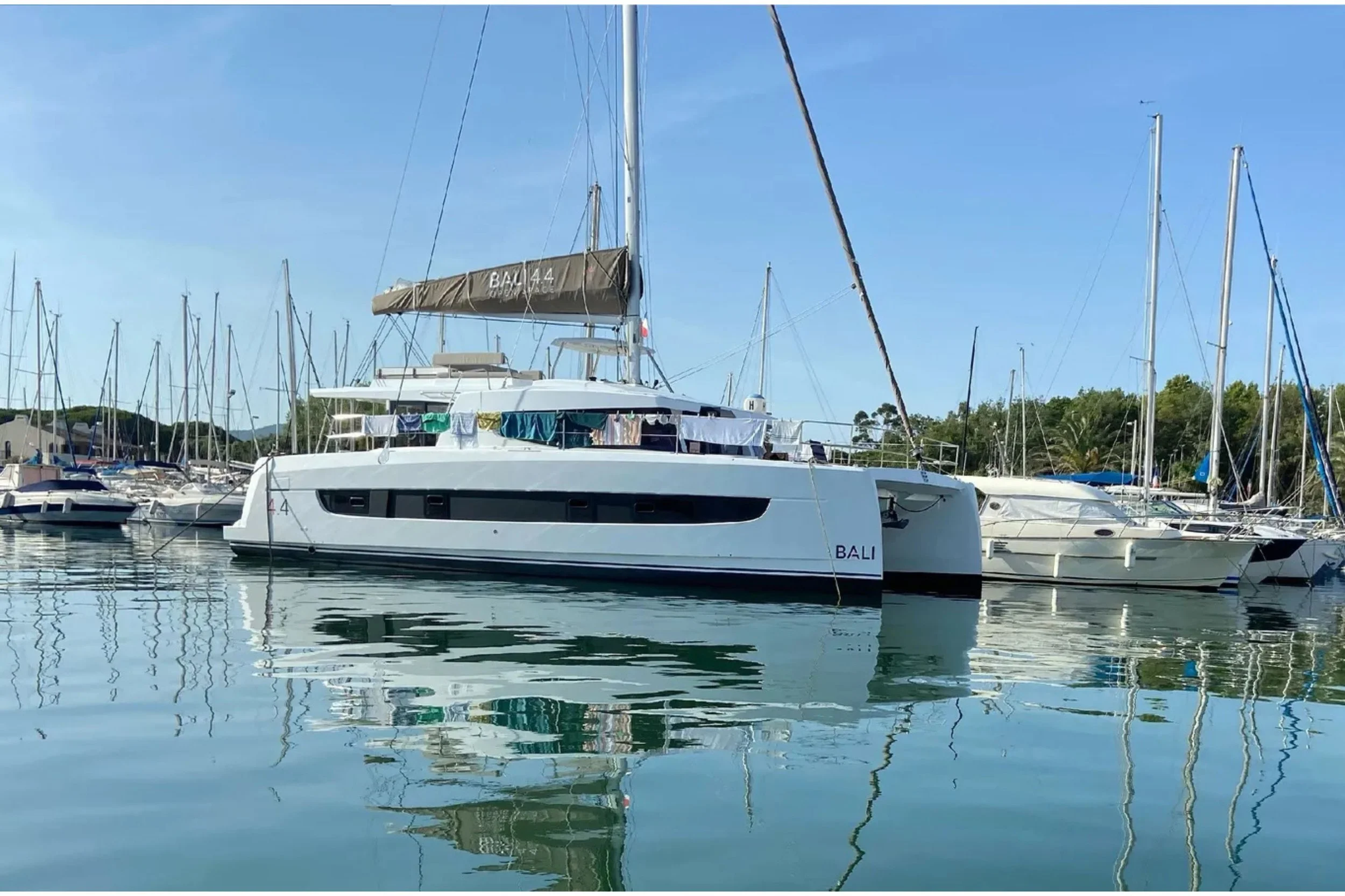 A large white yacht named BALI docked at a marina with multiple sailboats around it, calm water, and a clear blue sky.