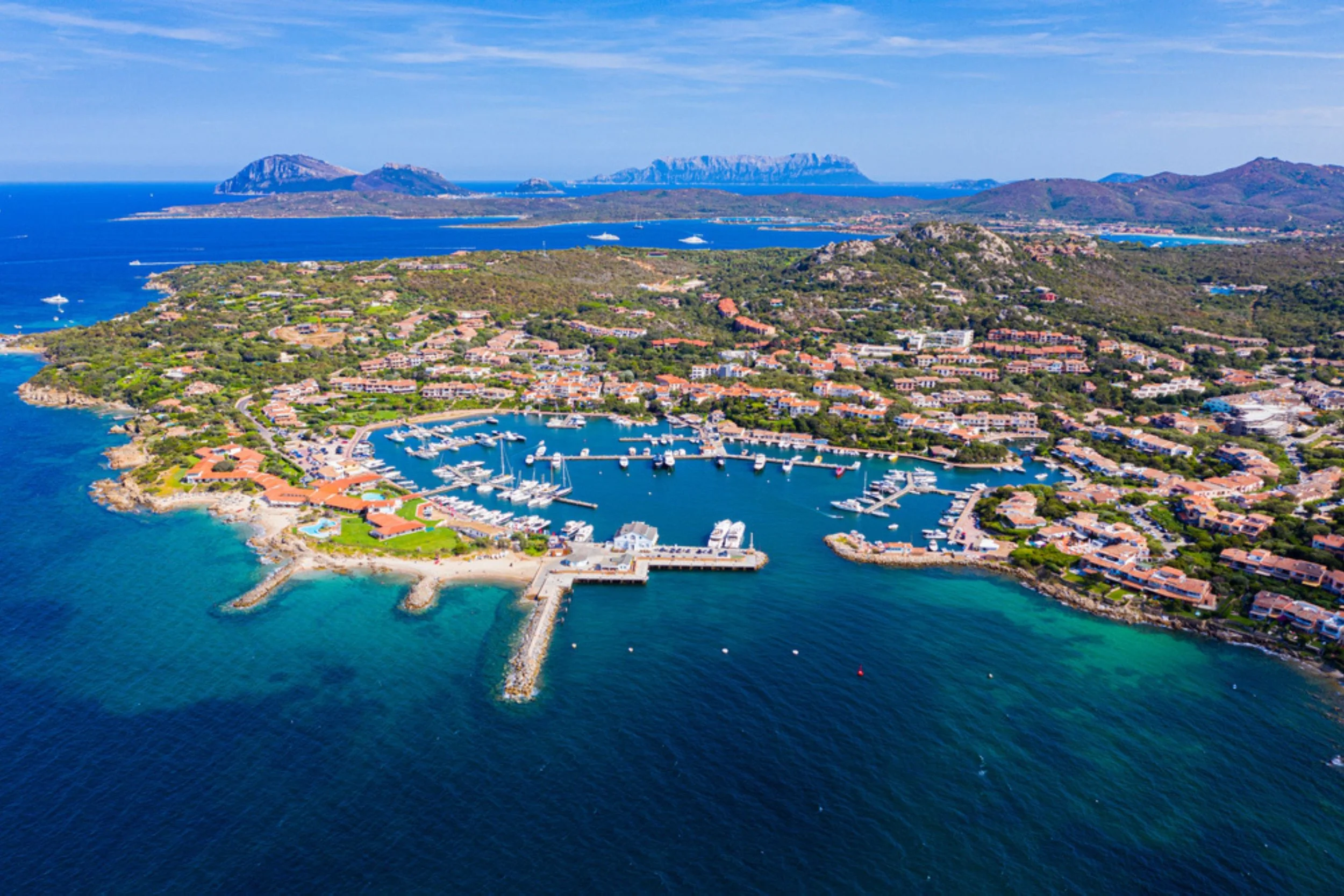 Aerial view of a coastal marina with sailboats and yachts, surrounded by residential buildings and green hills, with distant mountains and clear blue sky in the background.