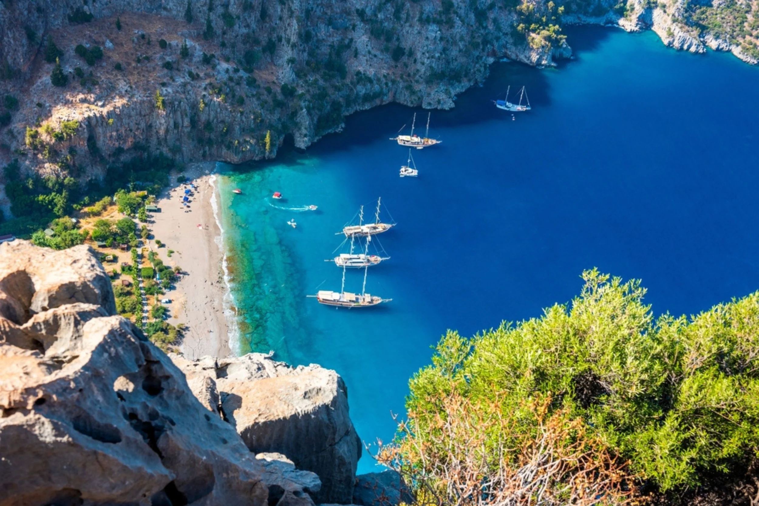 Aerial view of a bay with several sailboats anchored in clear blue water, a small beach with umbrellas and umbrellas, and green trees and rocky cliffs surrounding the bay.