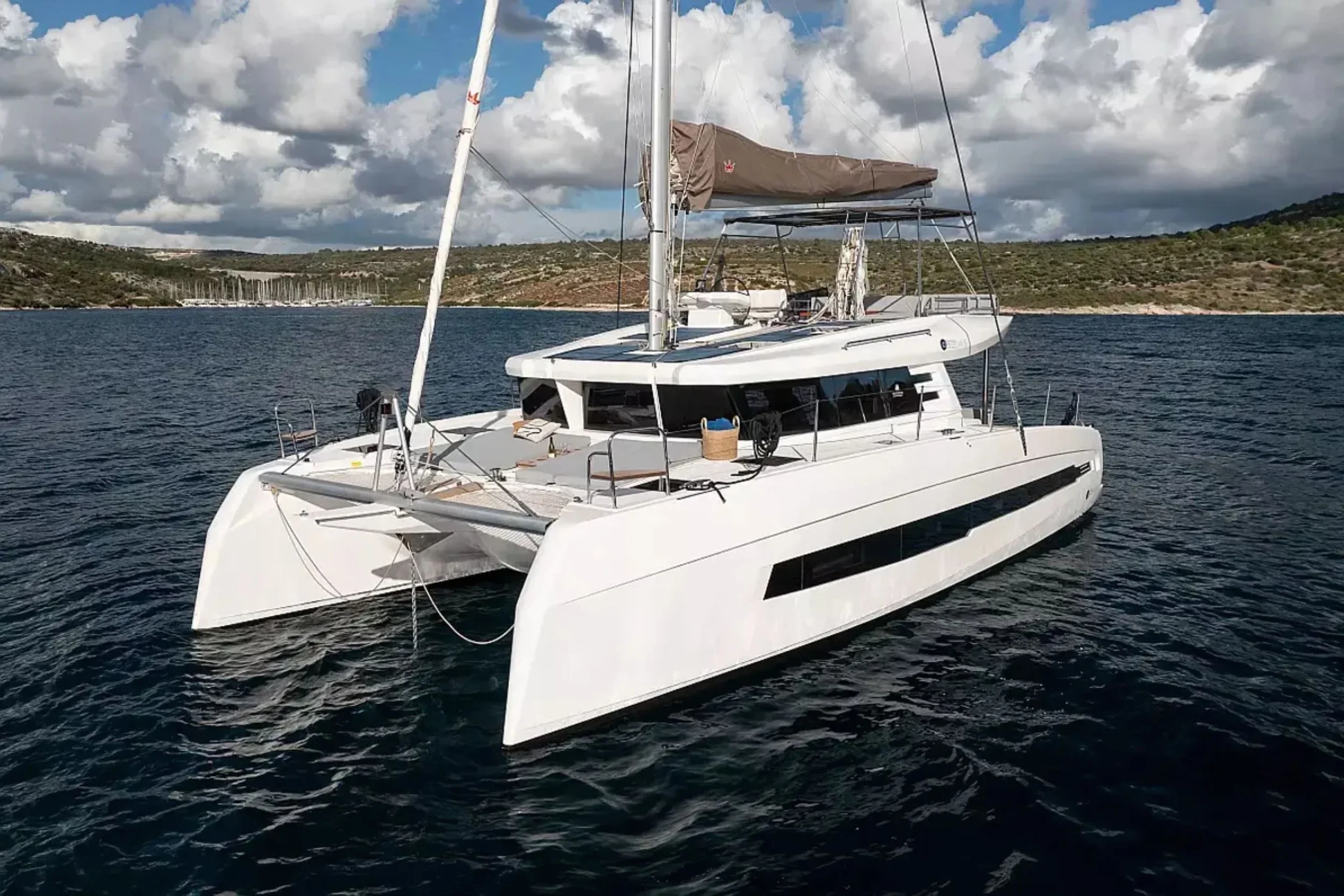 A white catamaran yacht floating on calm water with a landscape in the background, partly cloudy sky overhead.