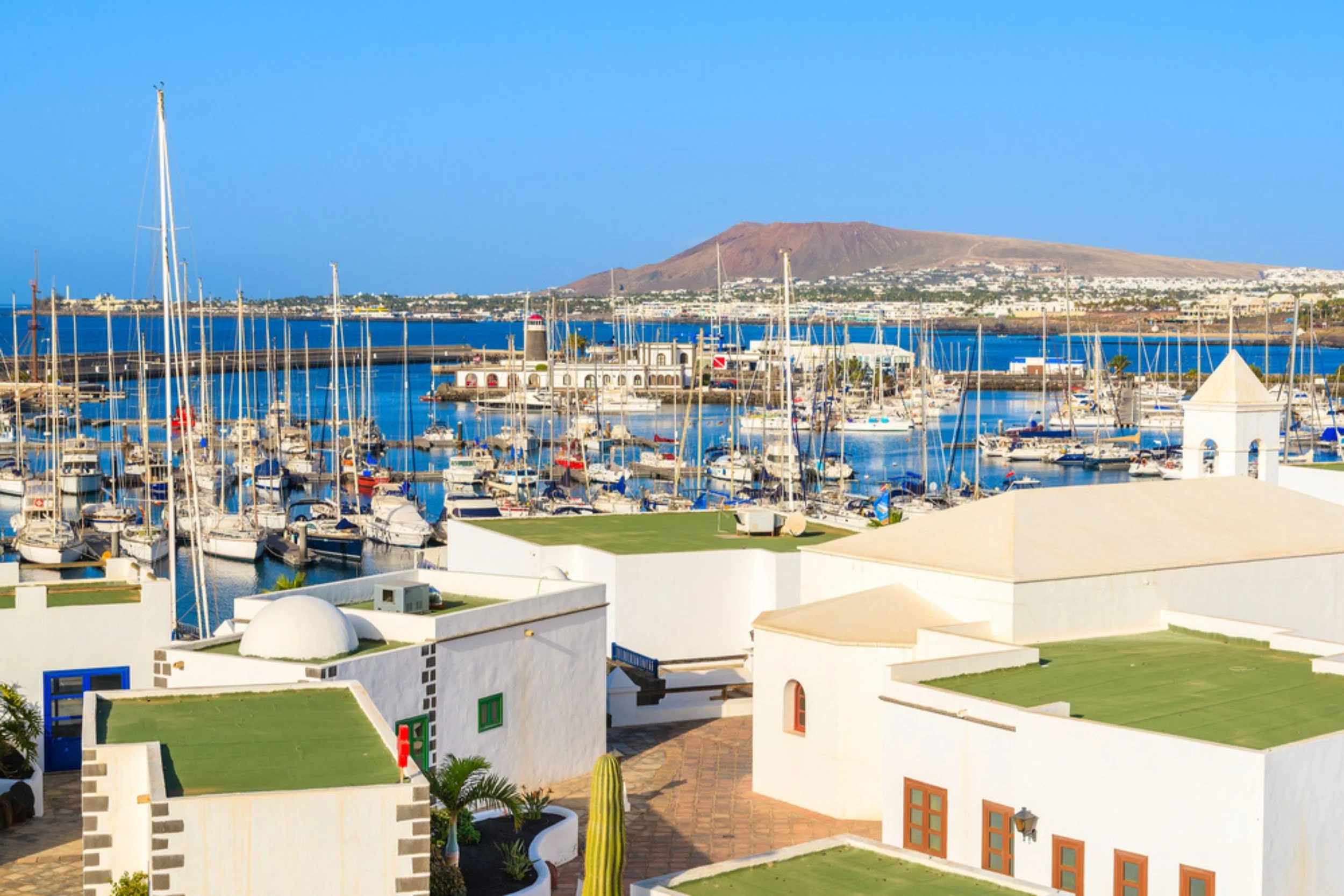 A marina with many sailboats docked, white buildings with green and brown windows in the foreground, and a mountainous island in the background under a clear blue sky.