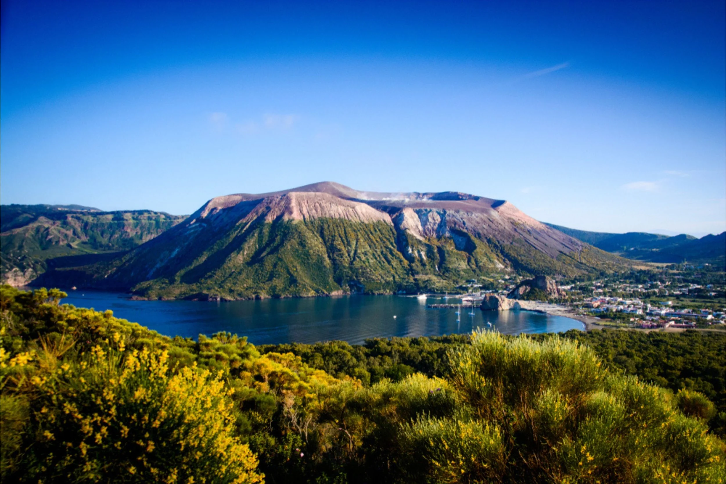 Scenic view of a large mountain with greenery, overlooking a bay with boats and a town along the shoreline under a clear blue sky.