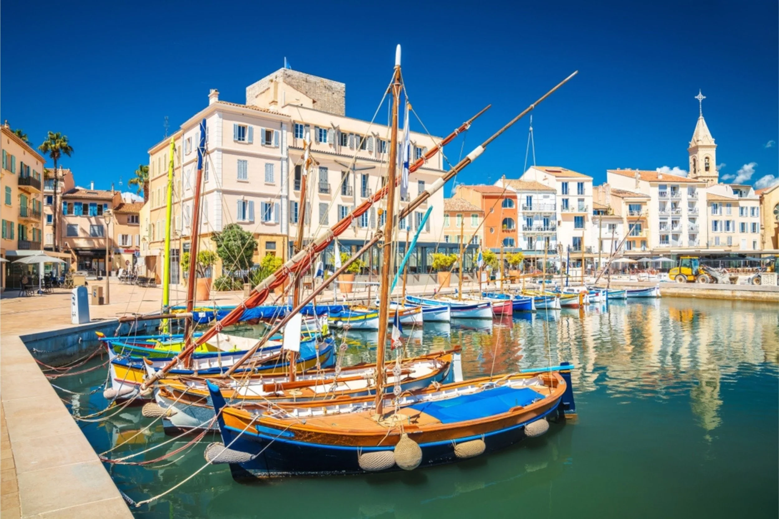 Boats docked in a harbor with colorful buildings and a church steeple in the background under a bright blue sky.