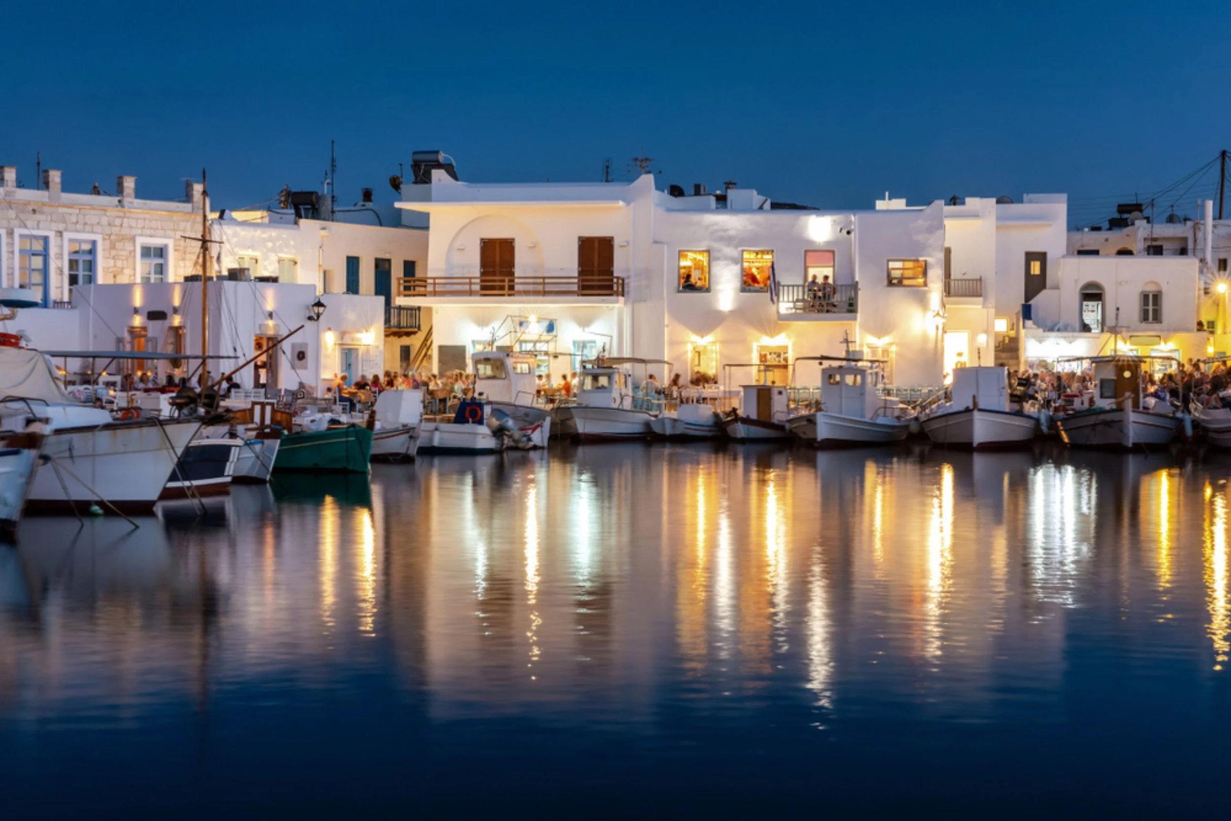 Night view of a marina with boats docked along the water, in front of white buildings with lit windows and outdoor dining, reflecting on the water.