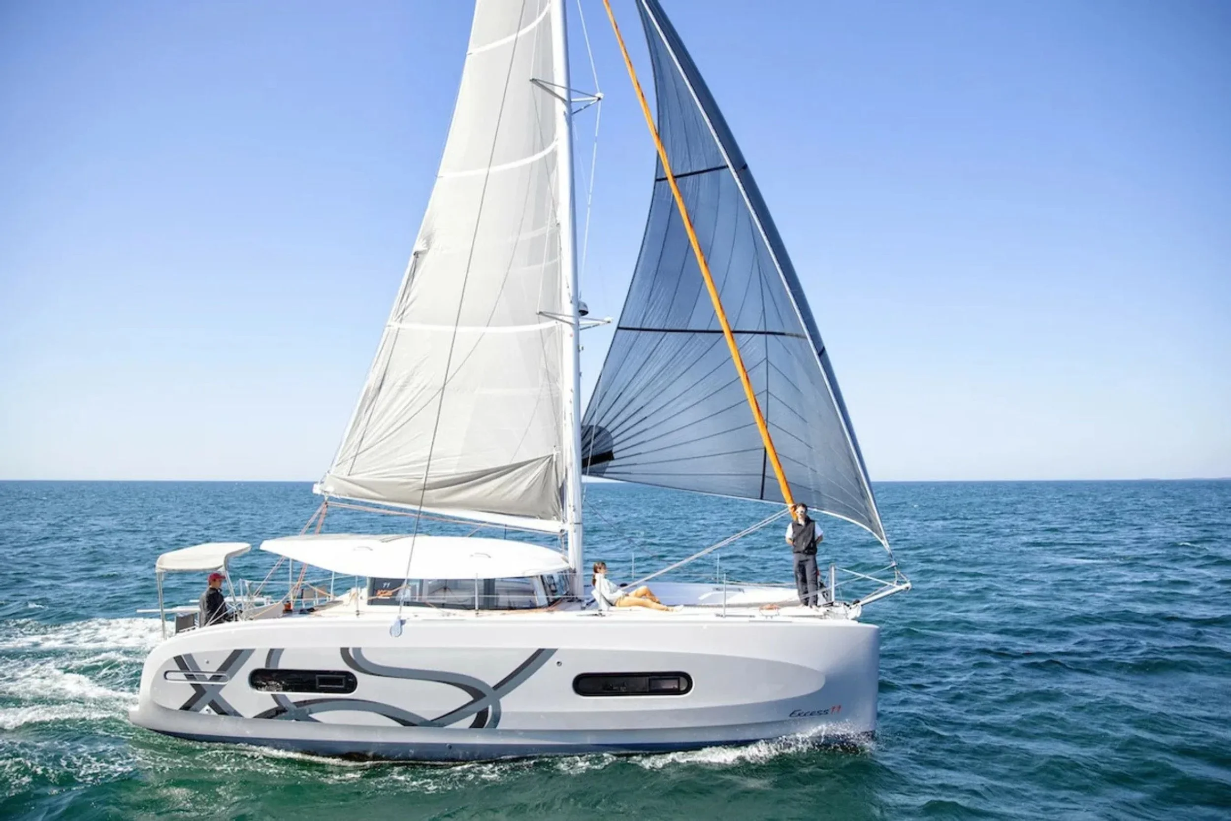 A white sailing yacht with a modern design and decorative black and gray patterns on its hull, sailing on calm blue ocean waters under clear blue sky. Three people are on board, one steering near the stern, one lying on the deck, and one standing near the bow adjusting the sails.
