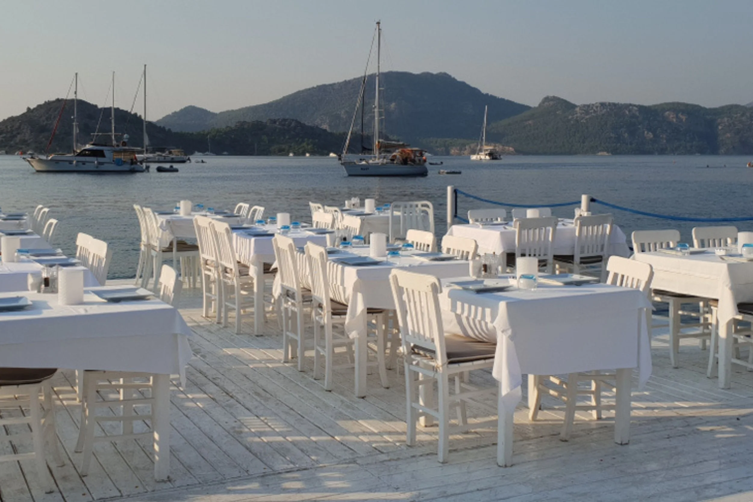 Outdoor dining area with white tables and chairs set on a deck by the water, with sailboats and small boats anchored in a calm bay, and distant rocky hills under a clear sky.
