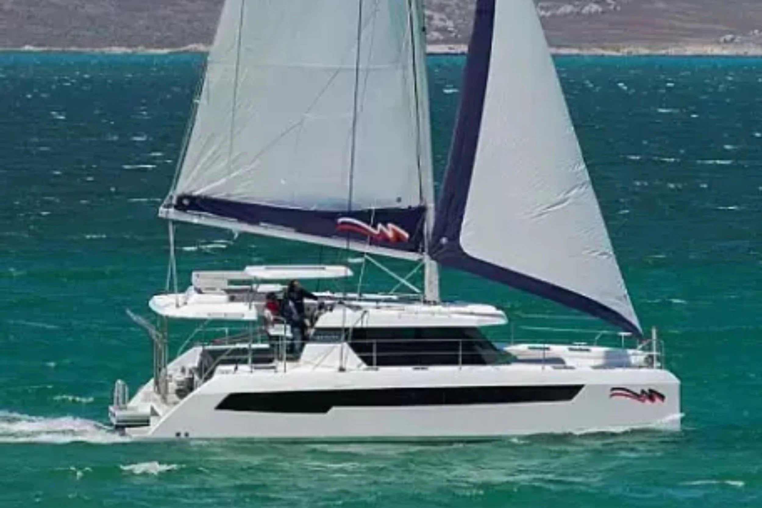 White sailing yacht with black accents and large sails on teal water, with a rocky shoreline in the background.