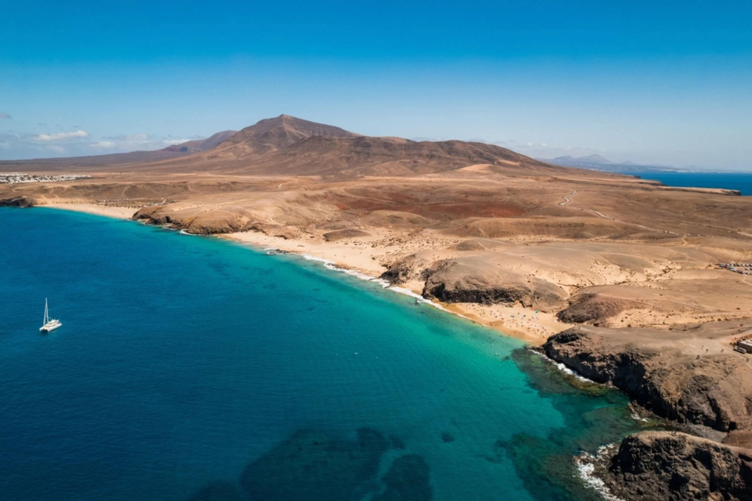 Aerial view of a coastal landscape with a beach, turquoise water, and arid hills and mountains in the background.
