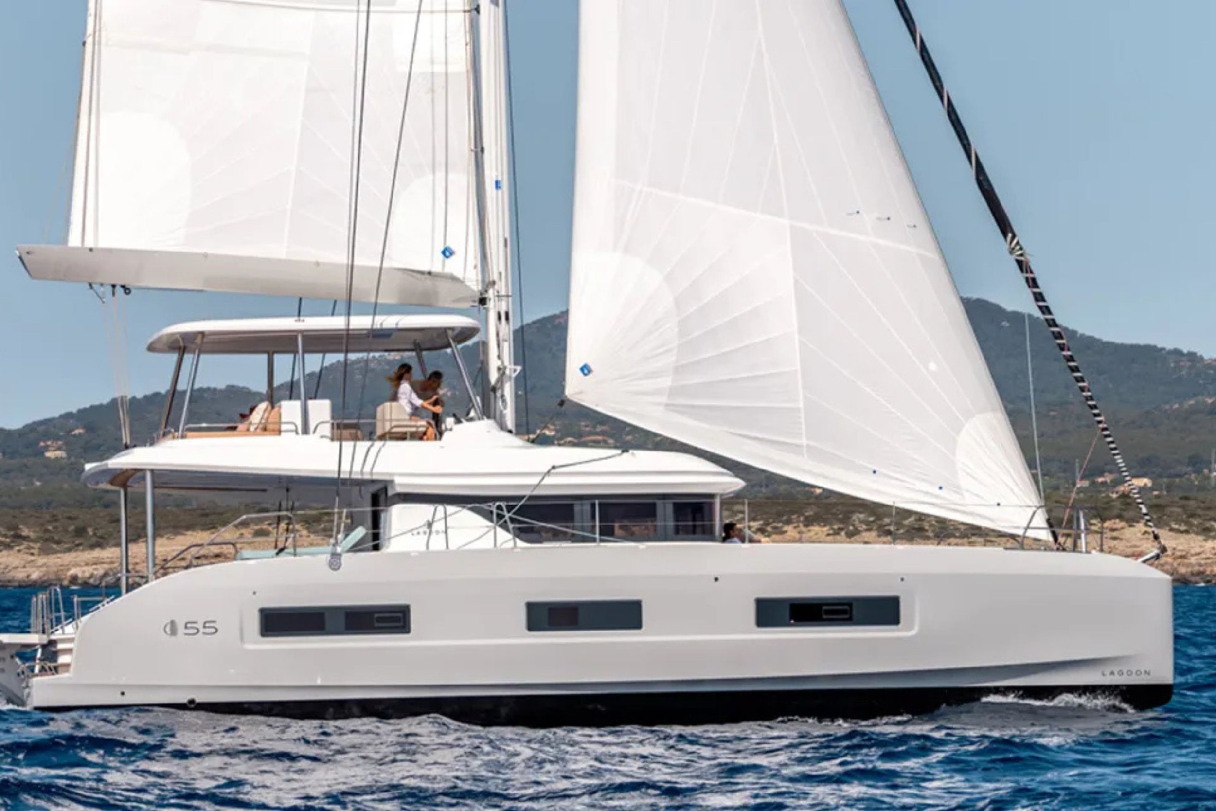 A white sailing yacht with large sails on the open water, with a hilly landscape in the background and two people sitting on the upper deck.