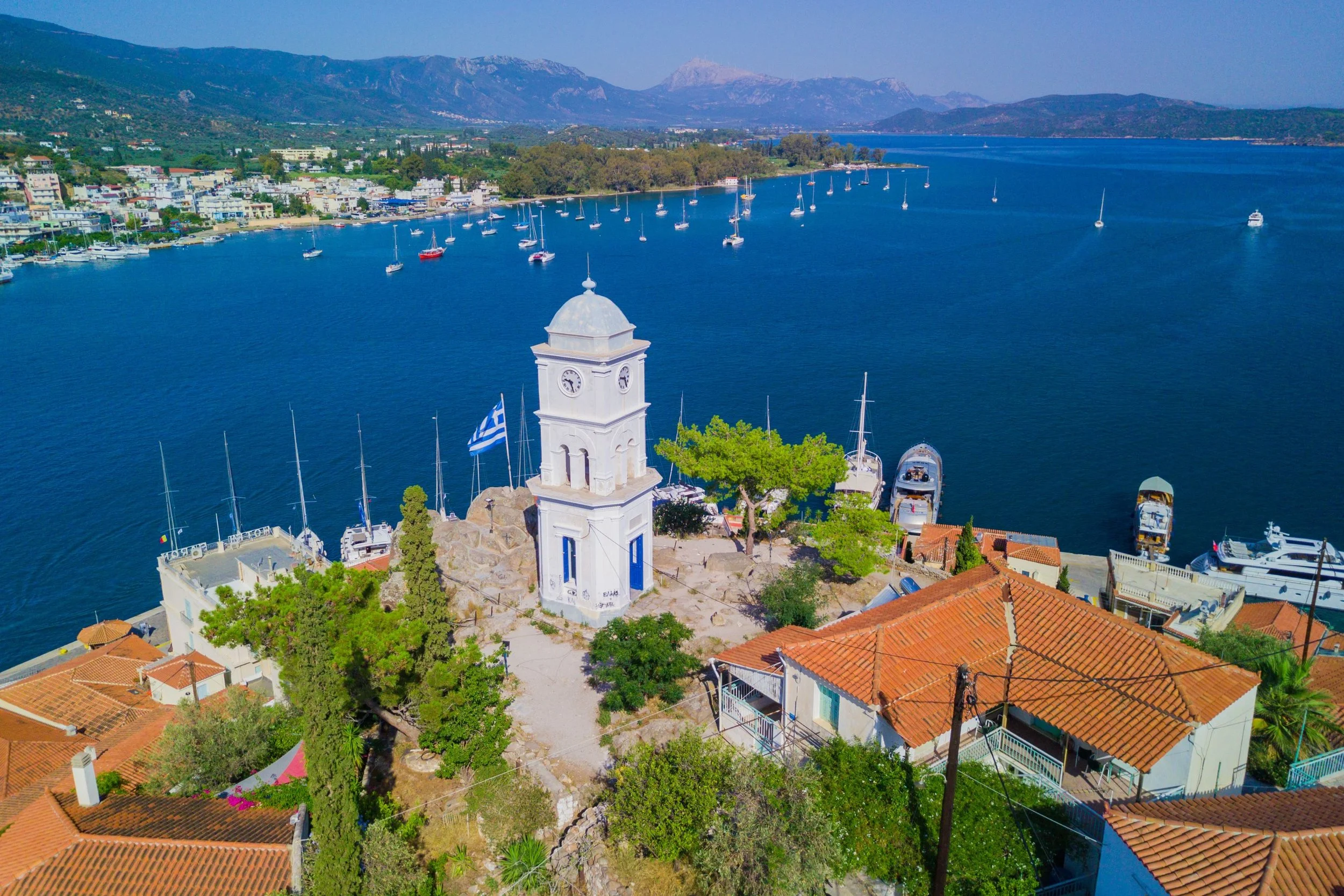A coastal town featuring a white clock tower with a blue door and Greek flag, orange-tiled roofs, and boats docked along the waterfront with mountains in the background.