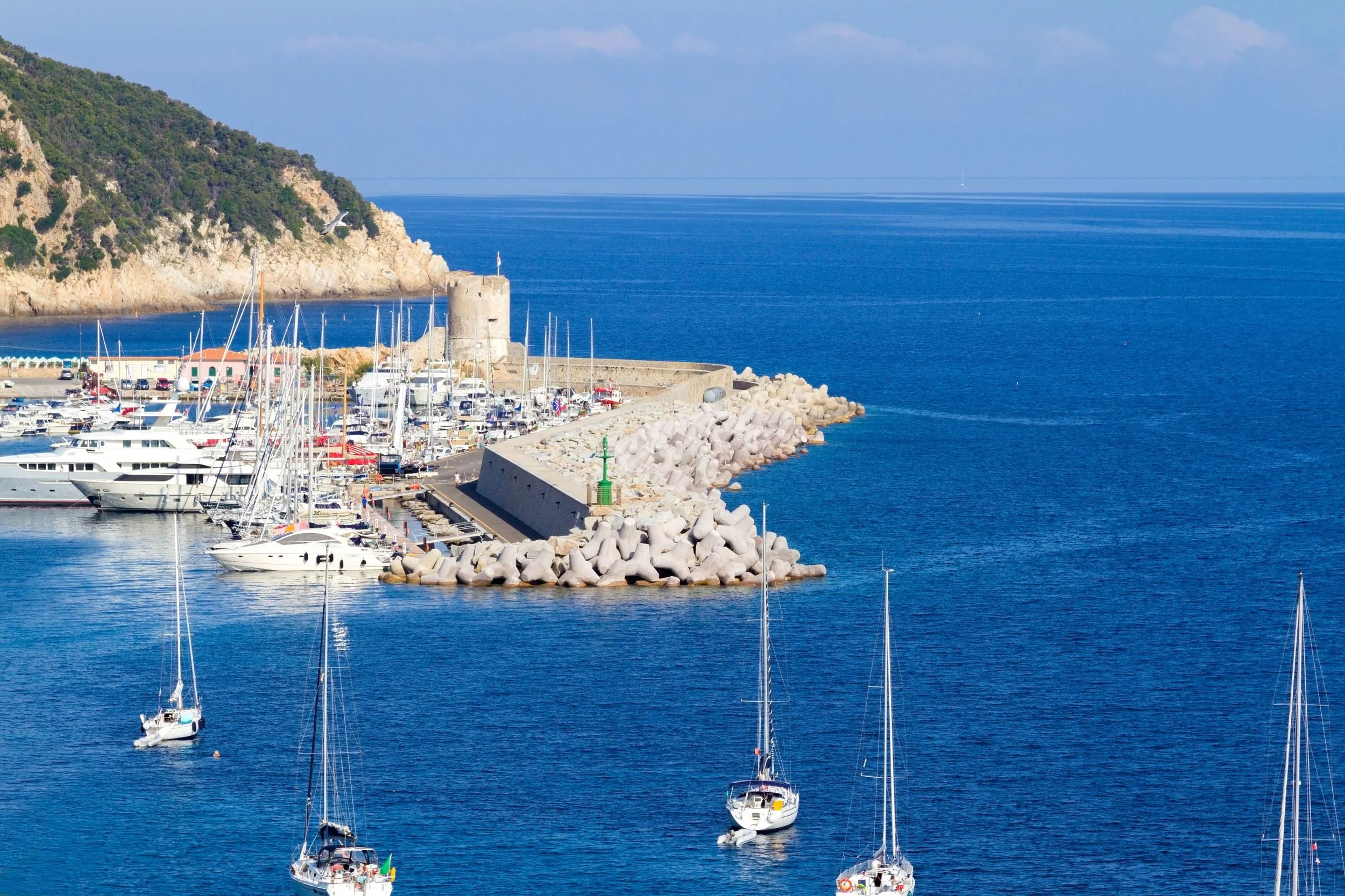 Marina with sailboats and yachts, stone breakwater with lighthouse, hill in background, bright blue sea and sky.