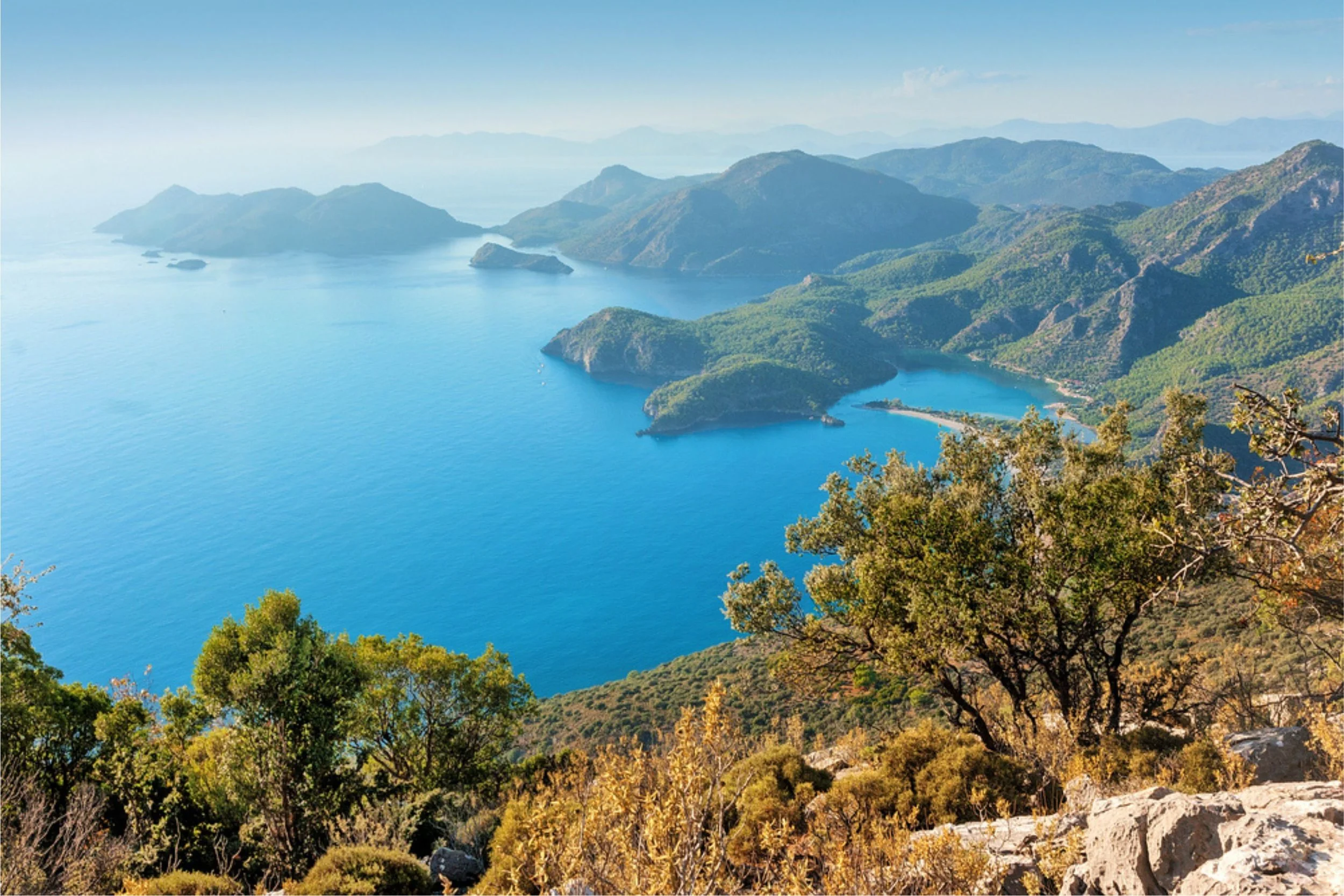 A scenic view of a large blue lake surrounded by green hills and mountains, with some tree foliage in the foreground.