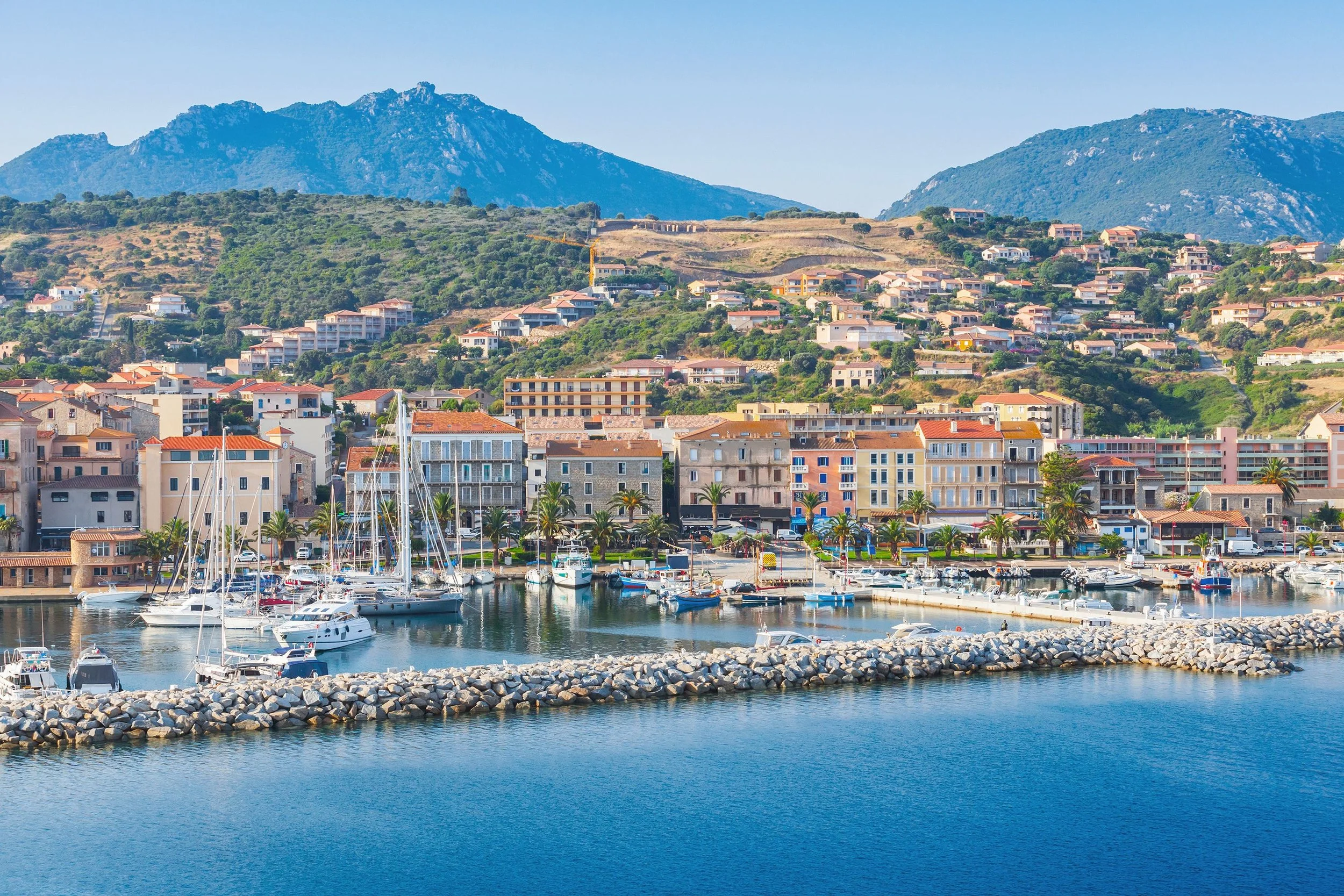A coastal town with sailboats docked in a marina, colorful buildings lining the waterfront, lush green hills in the background, and mountains under a blue sky.