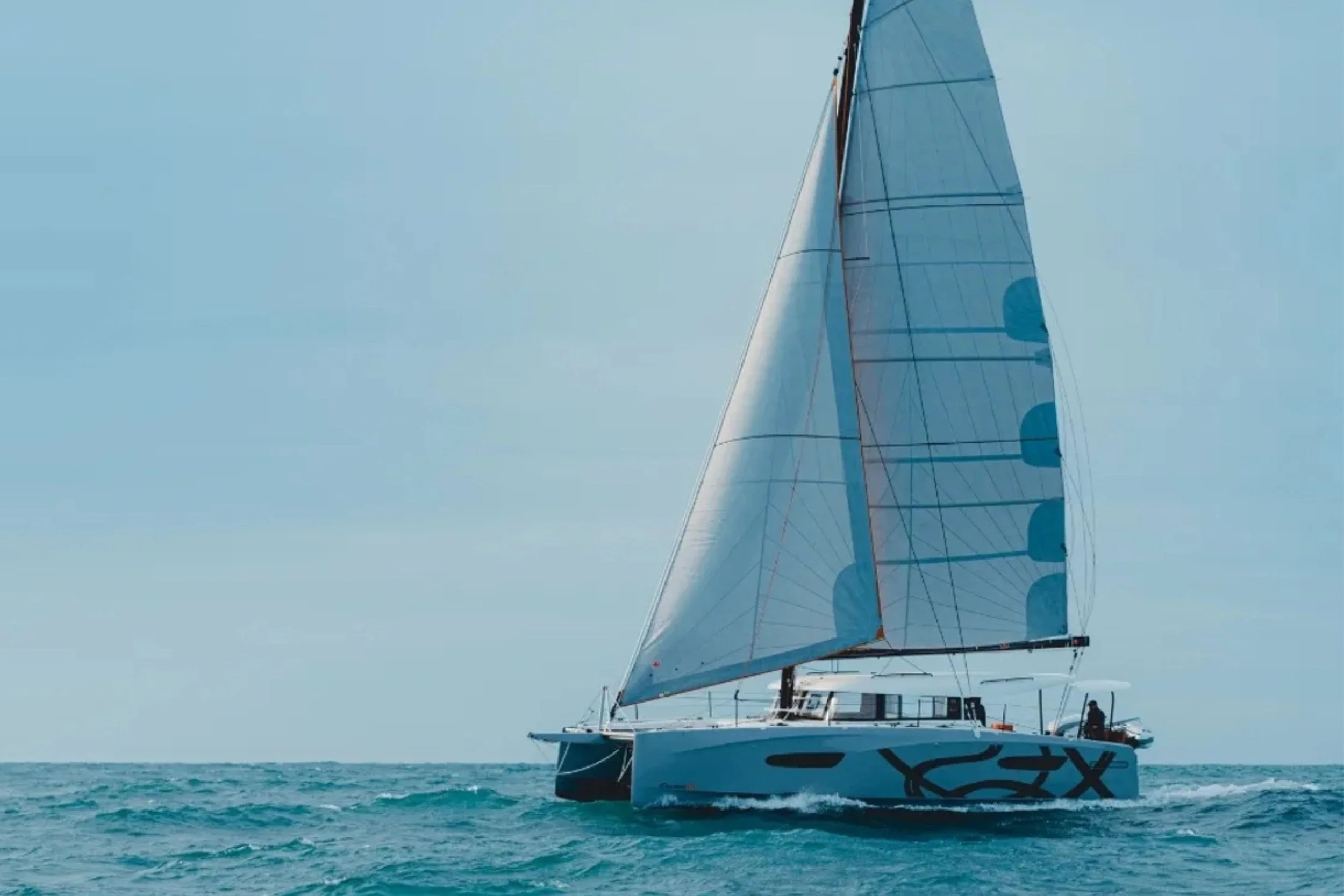 A sailboat with a large white and black hull and a tall white and blue sail on the ocean under a clear sky.