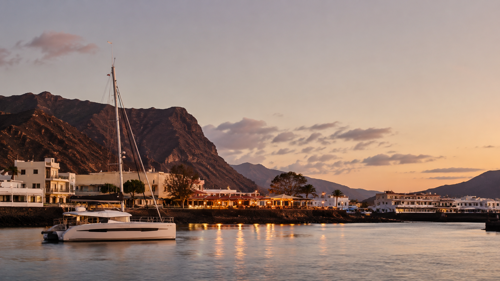 Modern catamaran at anchor near a peaceful harbour in the Canary Islands with softly lit waterfront restaurants at sunset