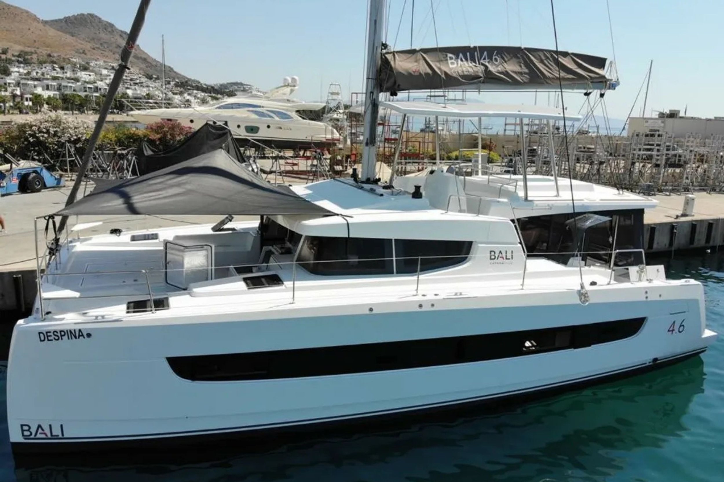 White sailboat docked at a marina with multiple other boats and yachts behind it, mountain in the background, clear sky, and calm water.