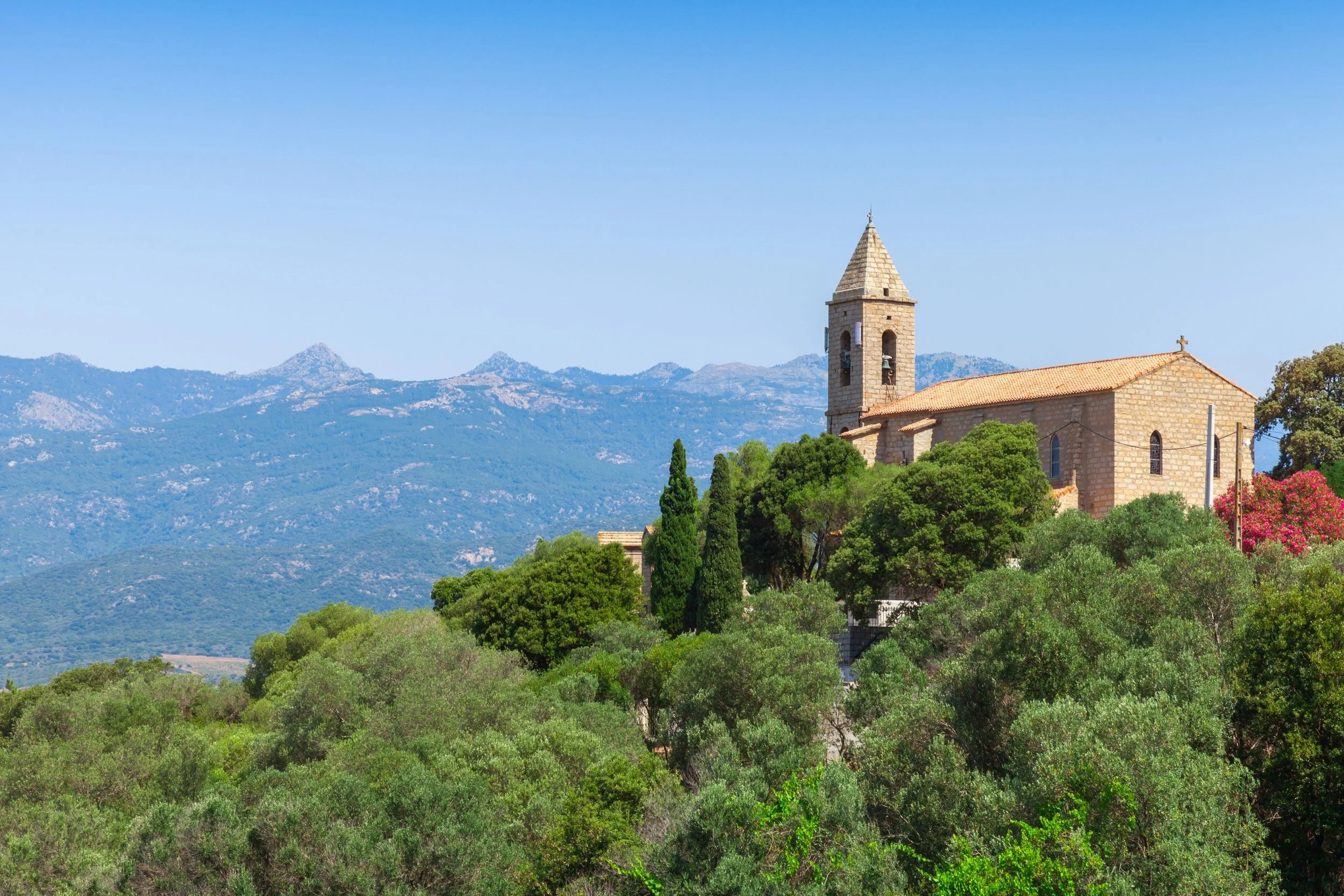 A stone church with a bell tower is situated on a hill covered with green trees, with mountain ridges in the background under a clear blue sky.
