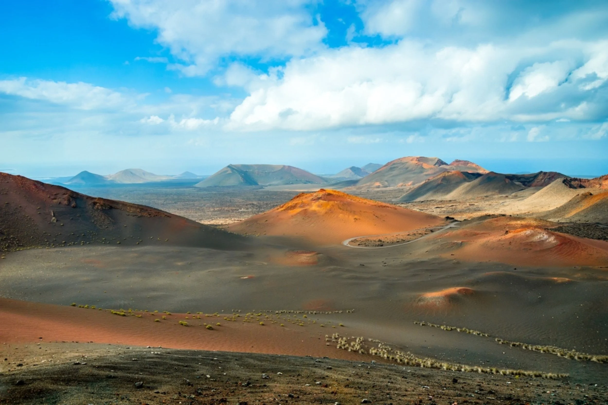 A vast volcanic landscape with multiple colorful volcanoes, some with reddish and orange hues, under a partly cloudy sky.