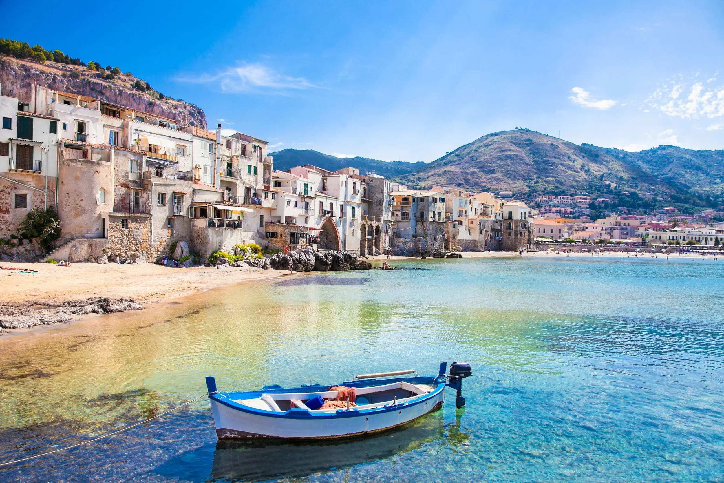 A small boat floating on clear blue water near a sandy beach with hillside and colorful buildings in the background.