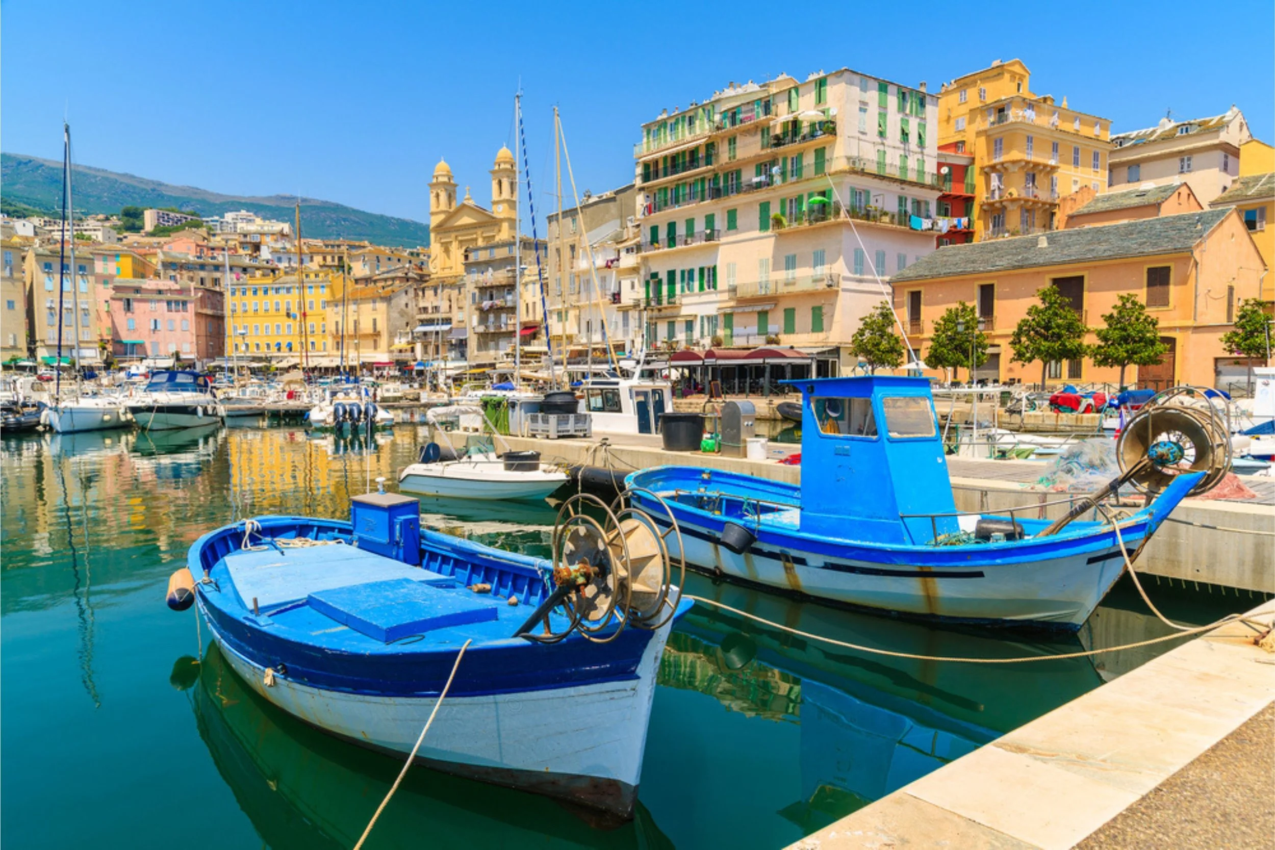 Colorful Mediterranean harbor with boats docked in calm water, surrounded by pastel buildings and a clear blue sky.