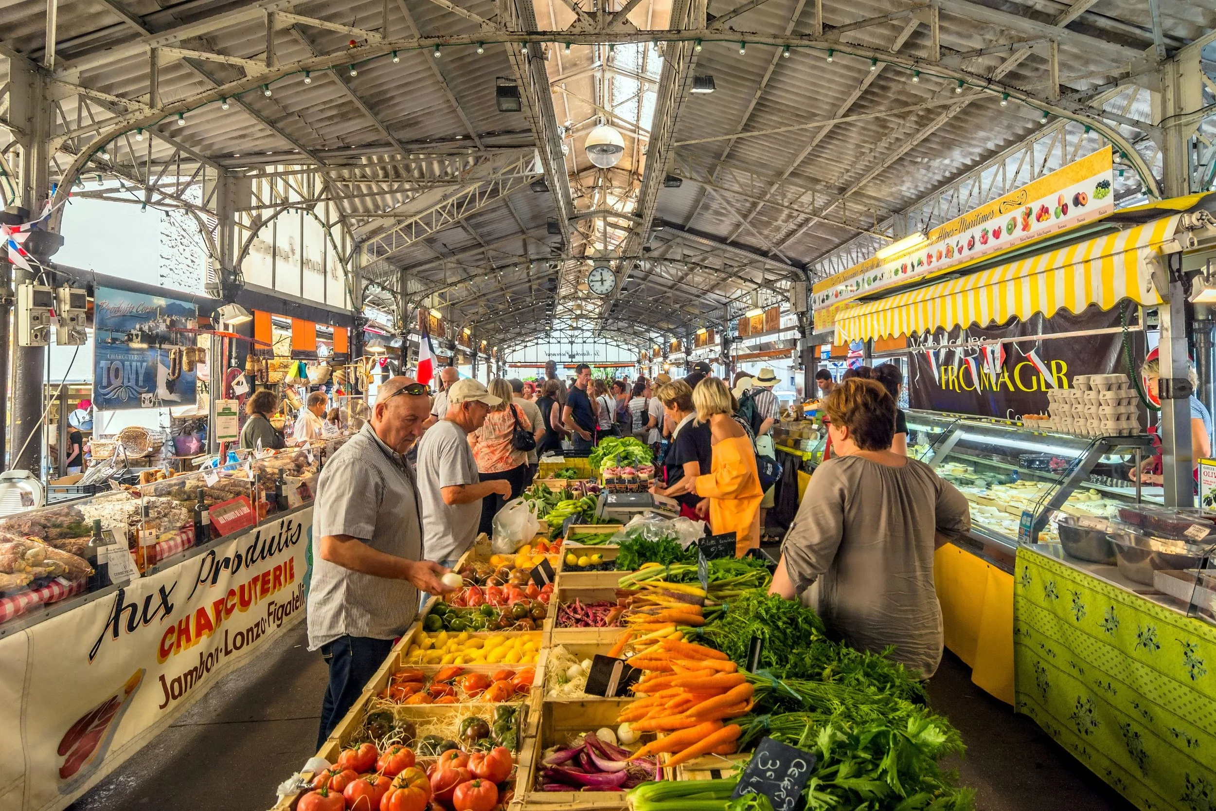 An indoor market with vendors selling fresh produce, including tomatoes, carrots, leafy greens, and other vegetables, under a high metal roof with hanging lights and a clock.