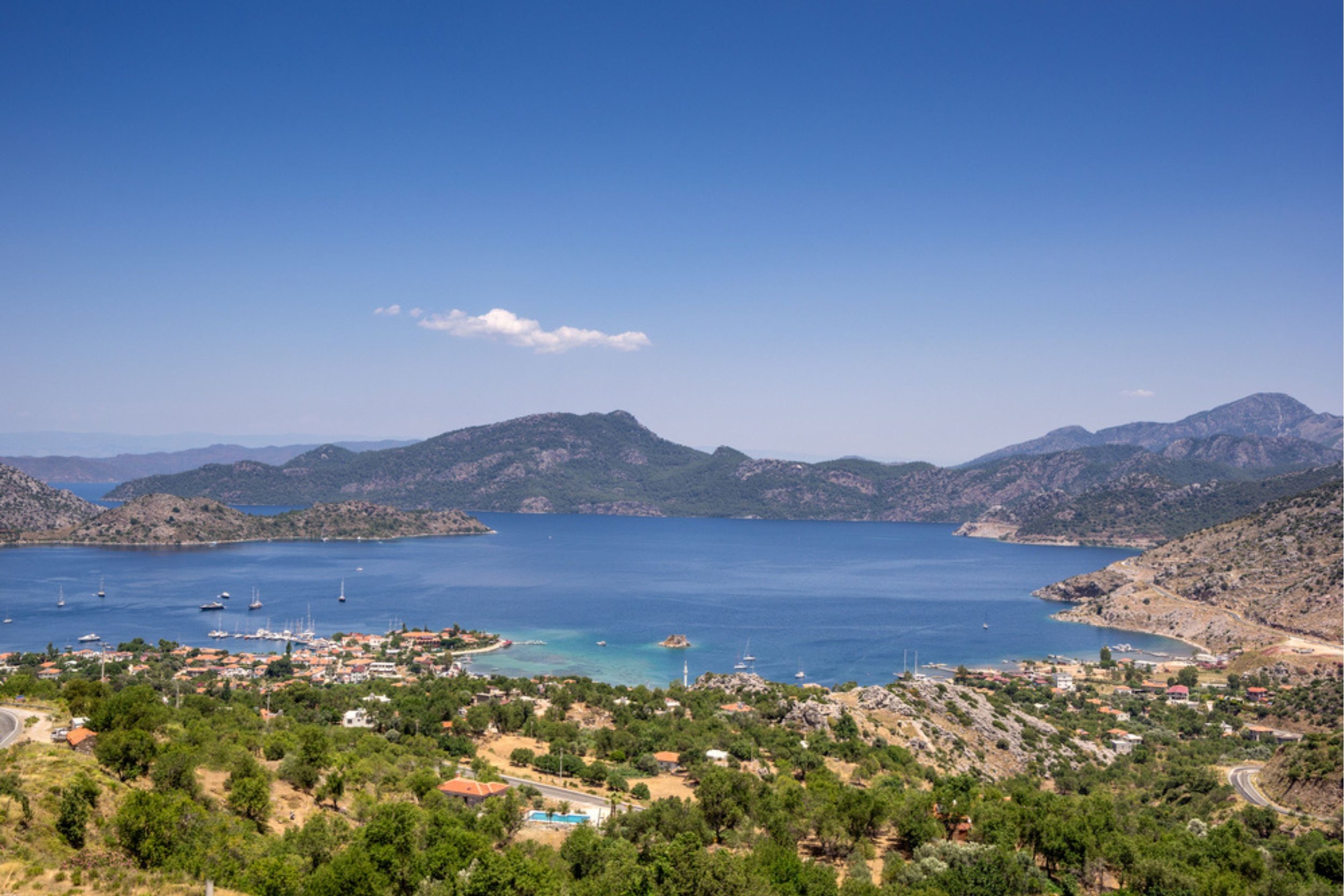 Landscape of a large blue lake surrounded by hills and mountains, with a small village and boats on the water under a clear blue sky.