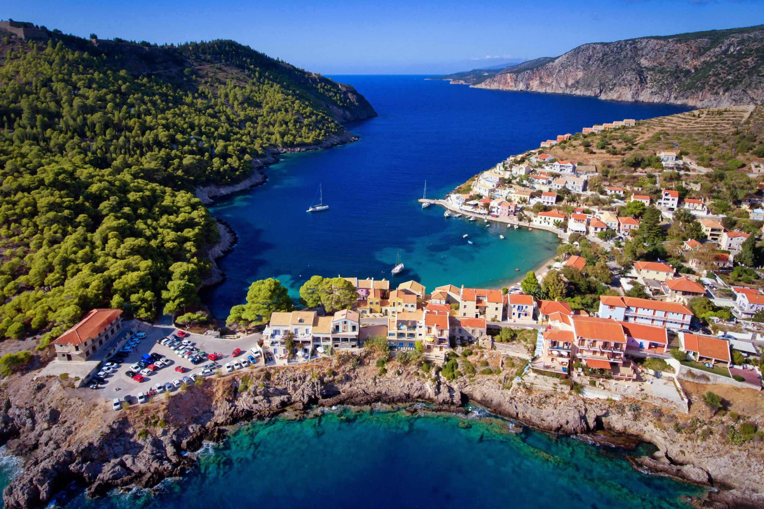 Aerial view of a coastal village with colorful houses, a small marina, and green hills surrounding a blue bay