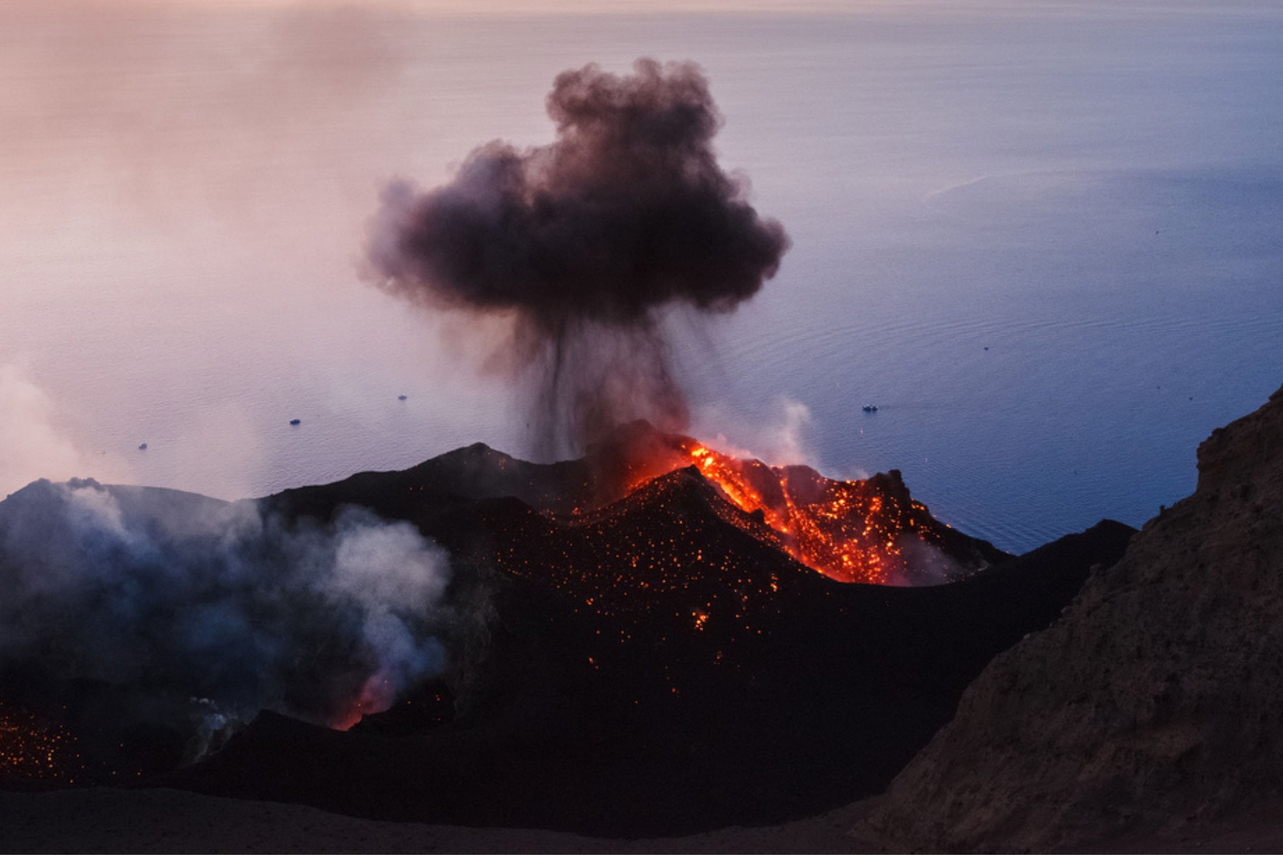 Erupting volcano with flowing lava, emitting black ash and smoke over dark mountains near a body of water at dusk.