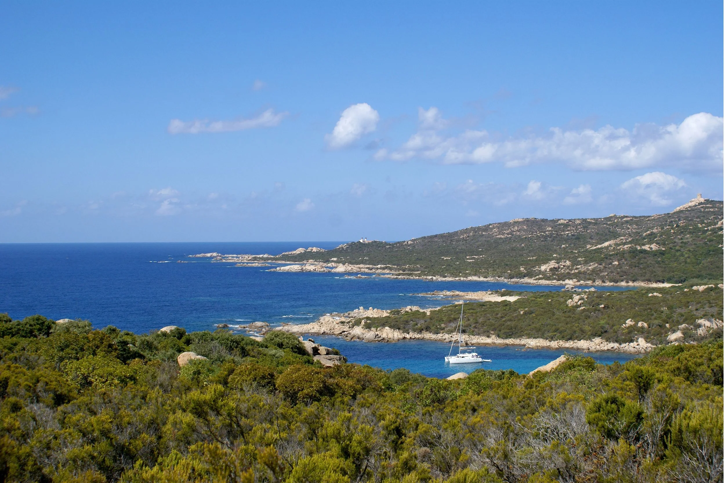A scenic coastal landscape with a blue sky and scattered clouds, a calm blue sea, rocky coastline, green shrubbery in the foreground, and a sailboat anchored near the shore.