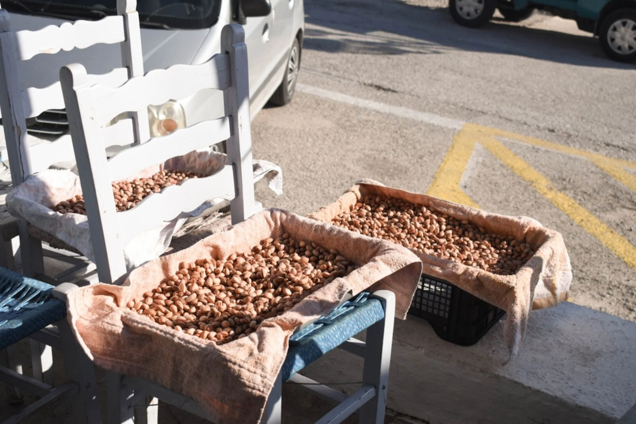 Street market stall with baskets of hazelnuts on white cloth and a white chair nearby, parked cars in background on asphalt road.