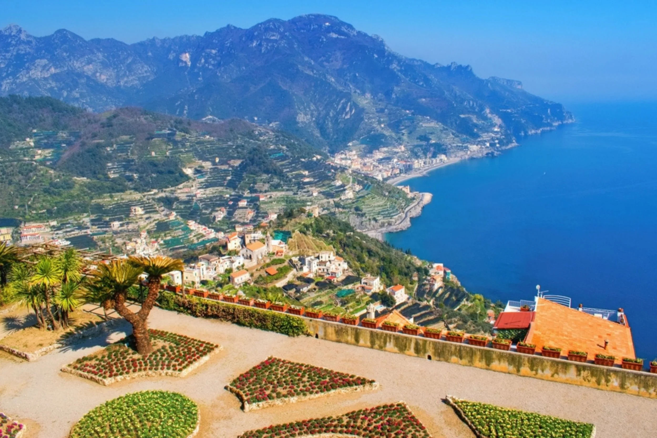 View of a coastal city with colorful buildings, terraced hillsides, and a deep blue sea, with a mountain range in the background.