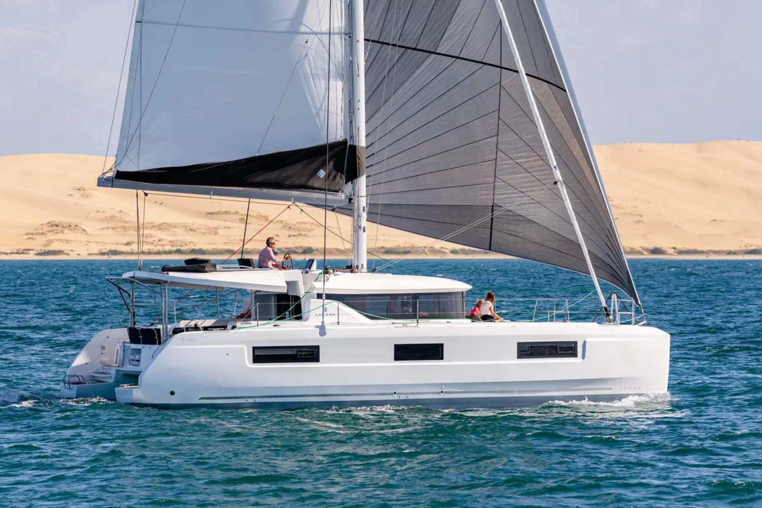 Sailboat on the water with two people on deck, yellow sand dunes in the background, blue ocean, gray sky, and large gray sails