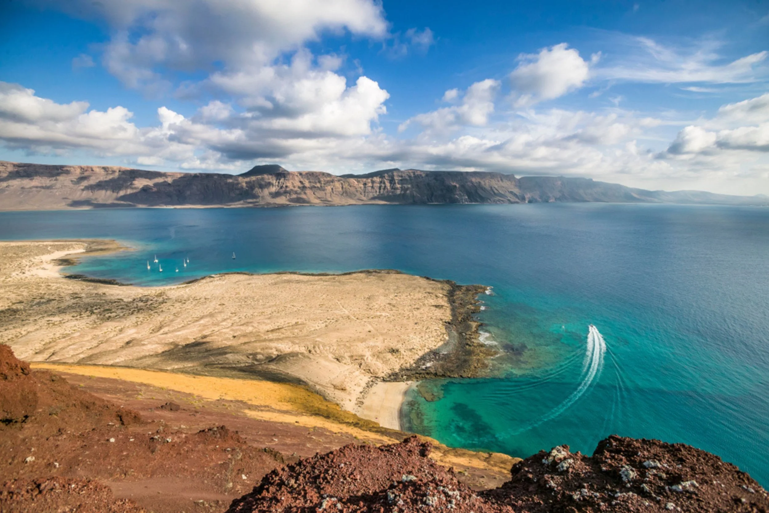 Aerial view of a coastal landscape with a sandy beach, turquoise water, and sailboats, surrounded by cliffs and mountains under a partly cloudy sky.